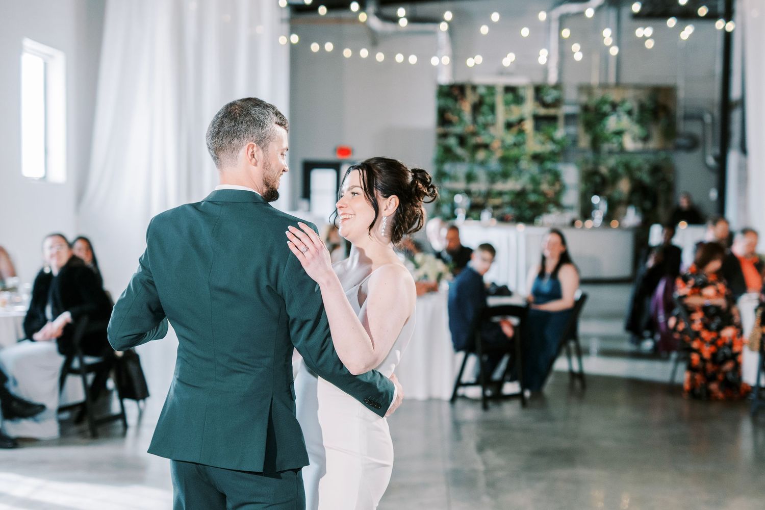Wedding couple dances in an industrial-style venue with string lights and greenery.