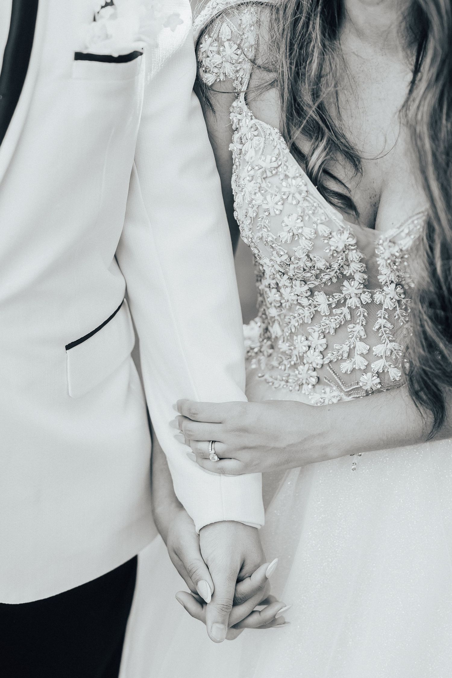 A close-up black and white photo showing hands holding between a white wedding dress and tuxedo jacket.