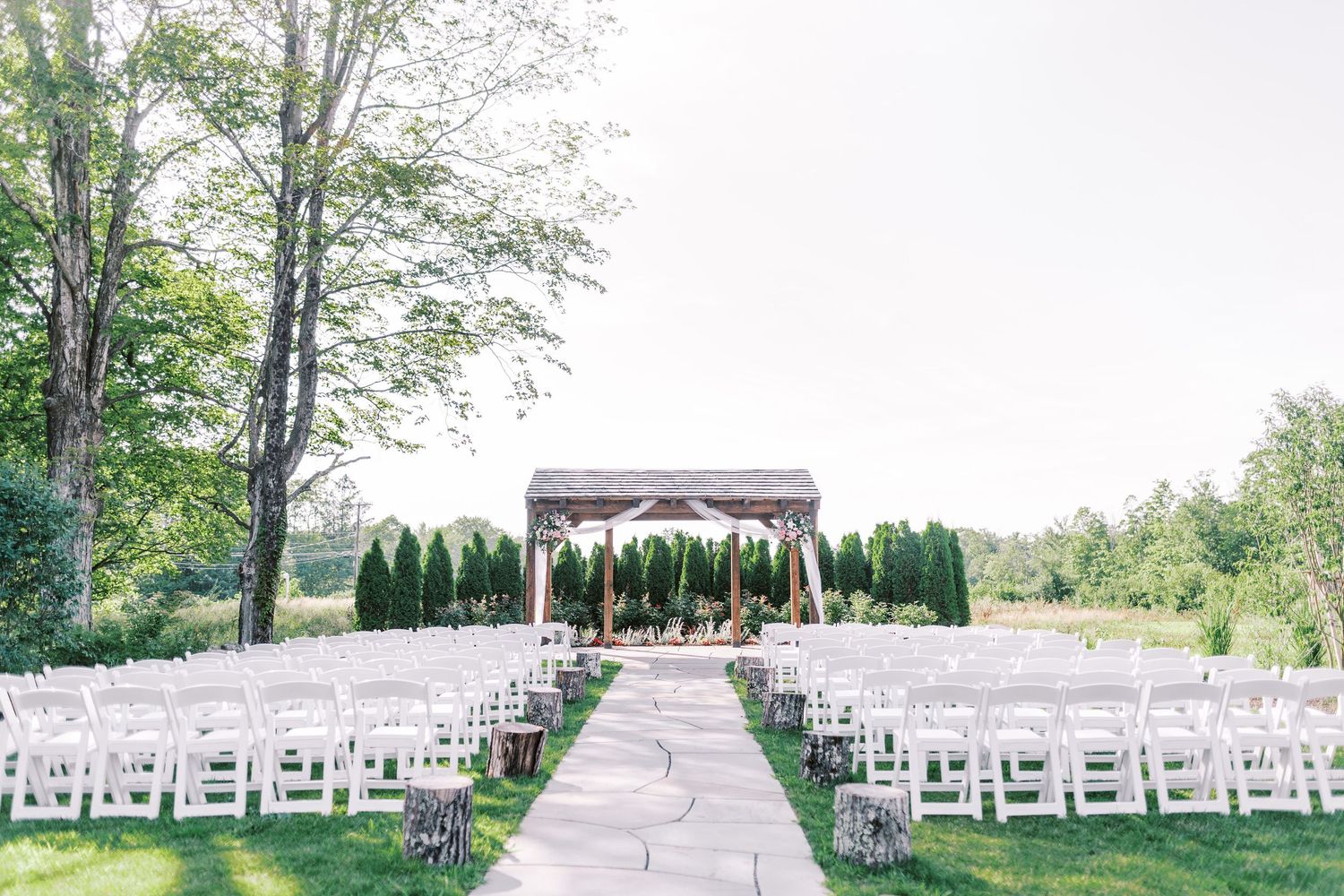Outdoor wedding ceremony setup with white chairs leading to a wooden pergola against a natural backdrop.