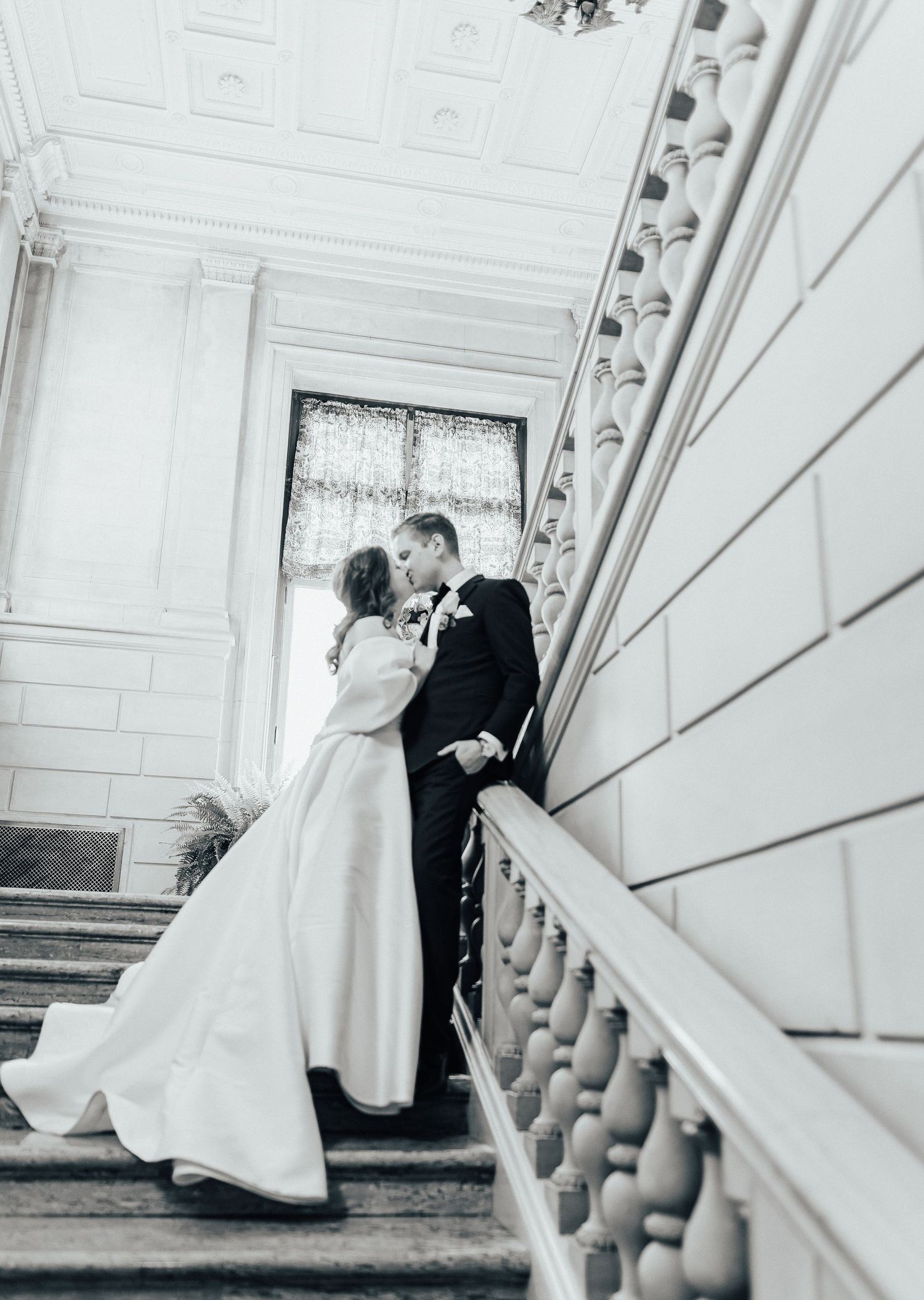 A black and white portrait of newlyweds kissing on an elegant staircase with ornate architectural details.
