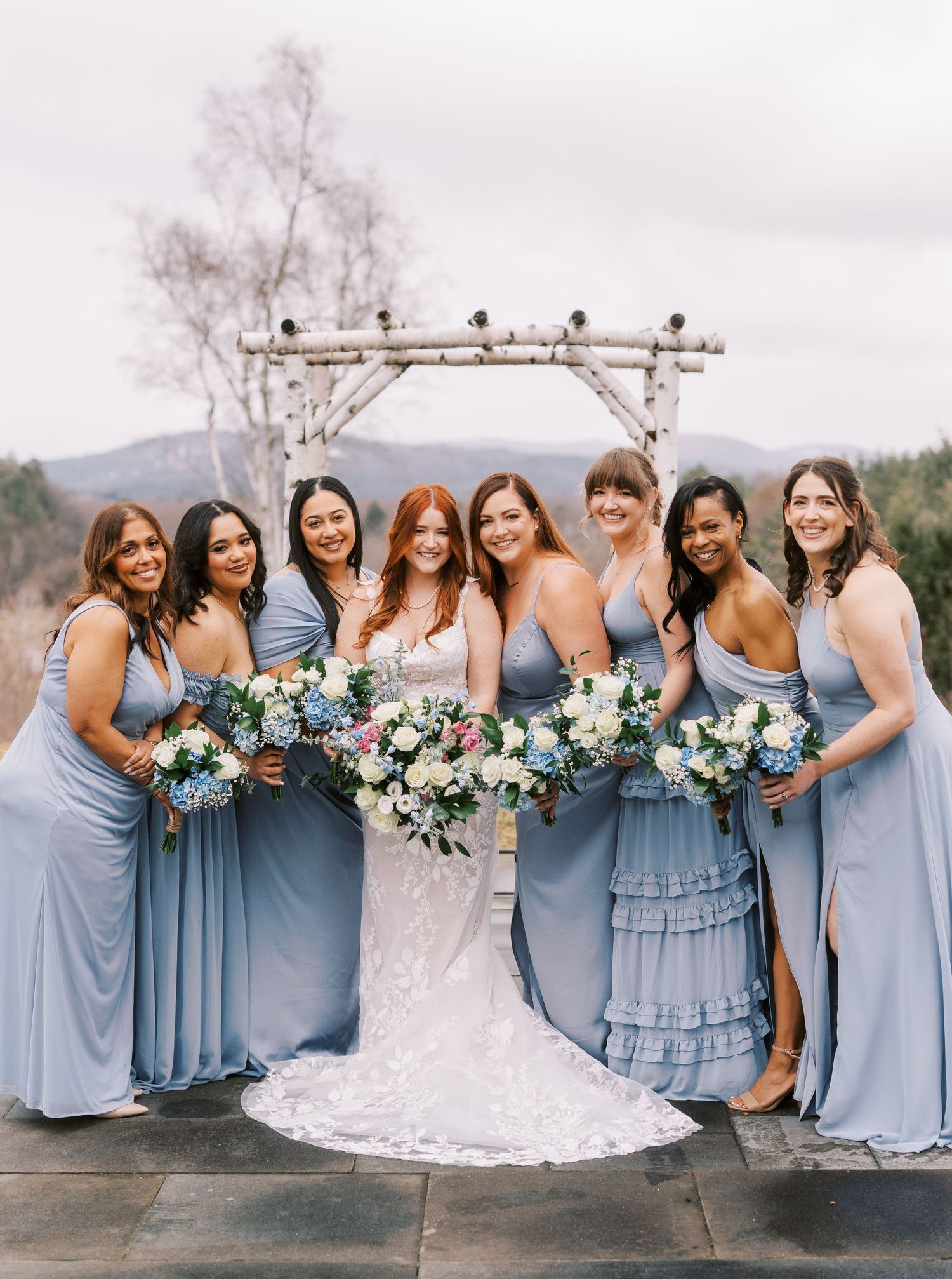 Bridesmaids in flowing dusty blue dresses pose together with colorful bouquets under a rustic outdoor arch.