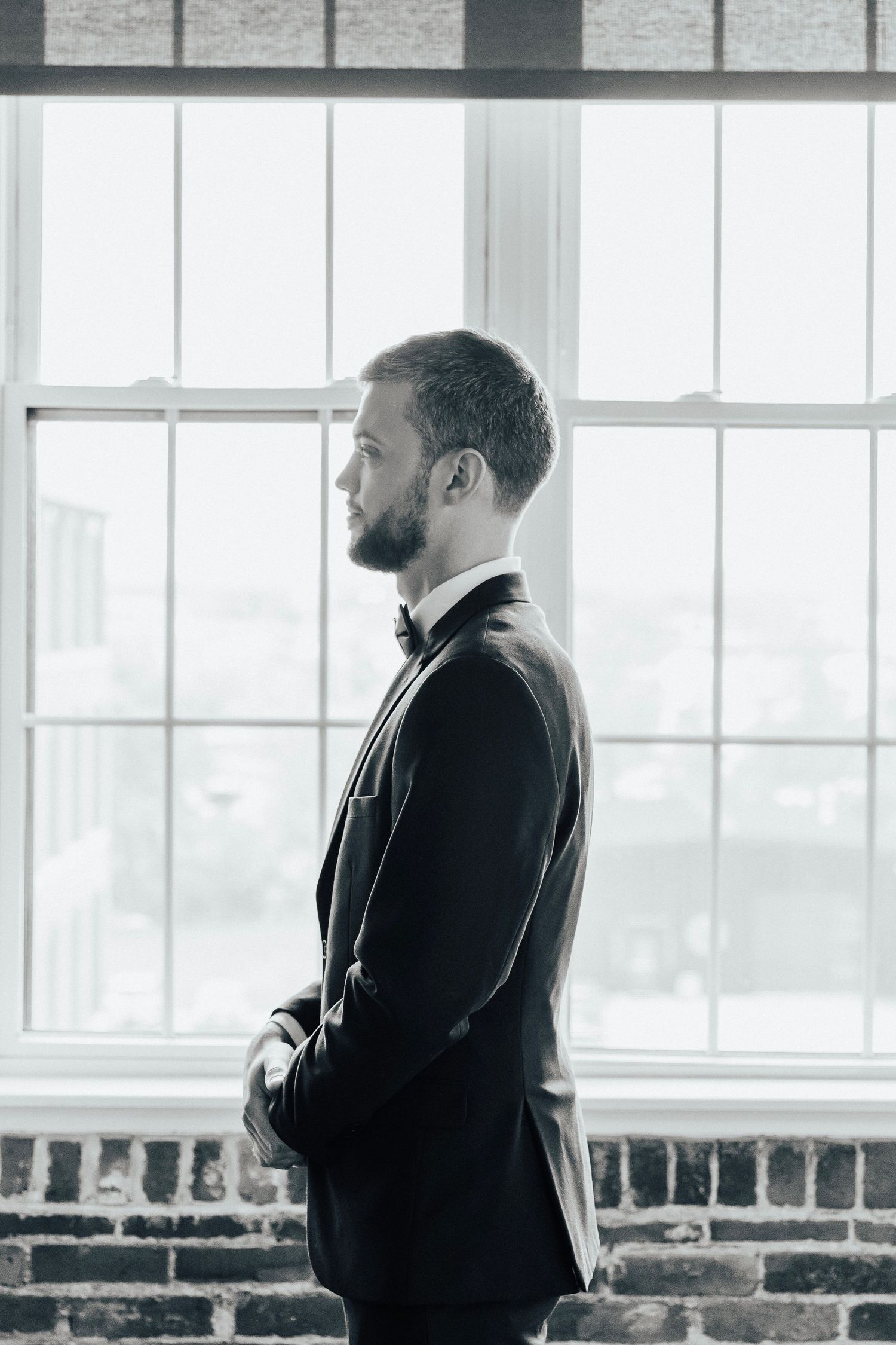 Formal portrait of groom in black tuxedo standing by industrial windows with exposed brick.