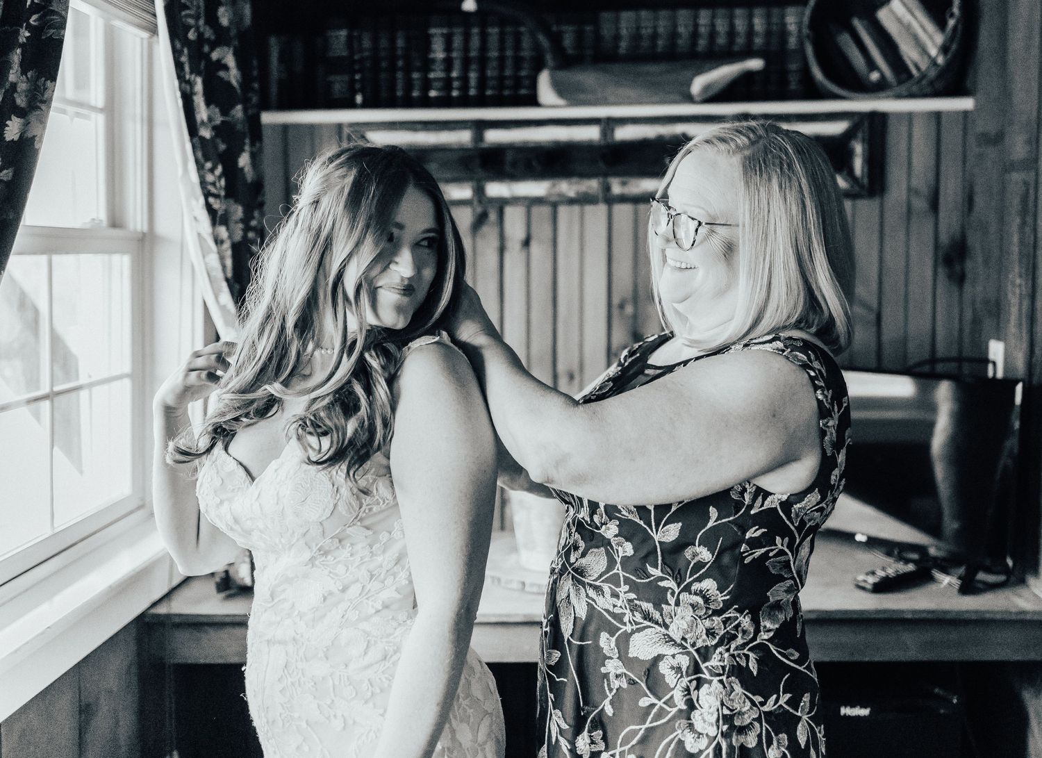 A black and white candid moment captured during wedding preparations by a window.