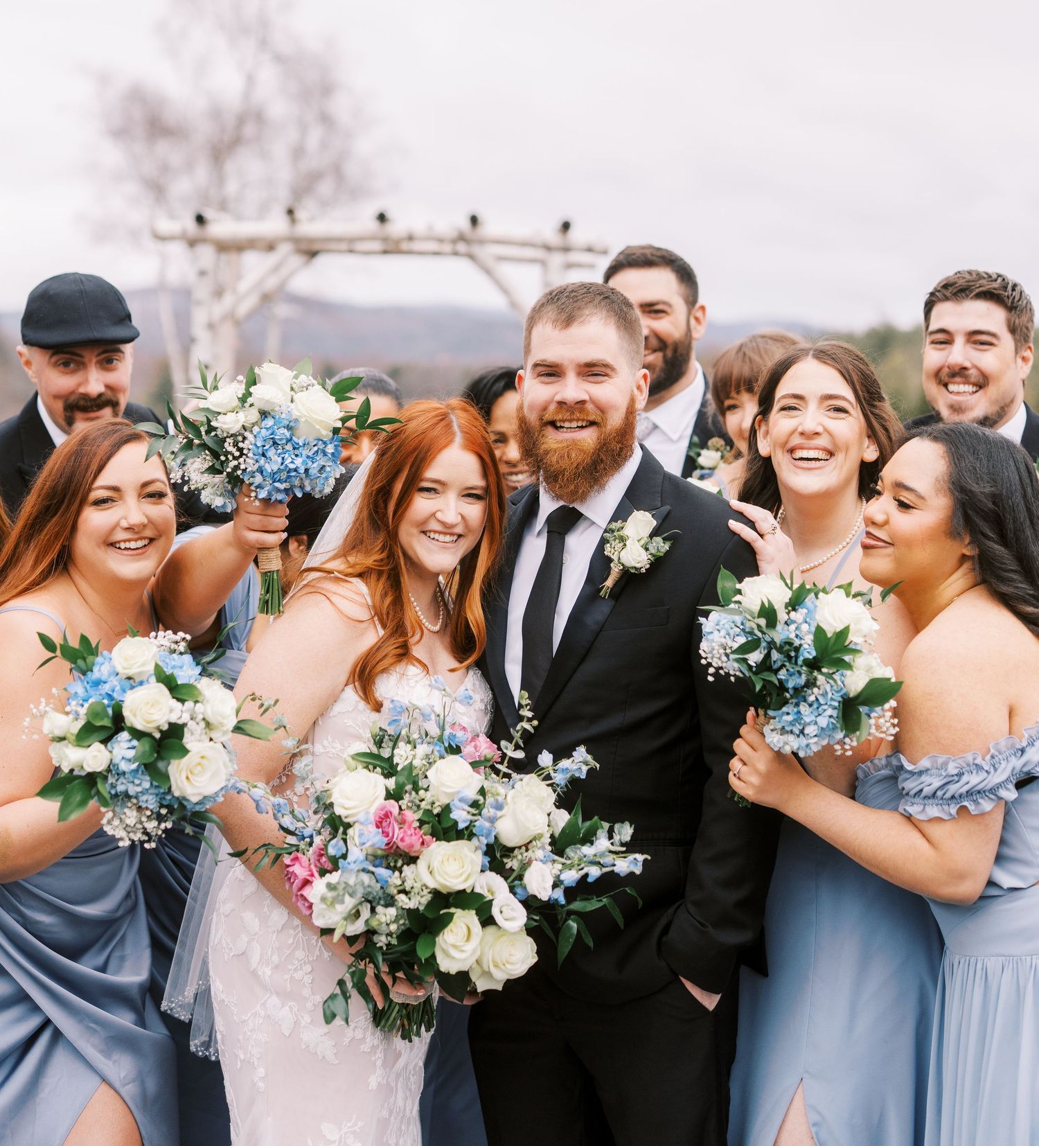 A joyful wedding party with bridesmaids in blue dresses holding colorful bouquets outdoors.