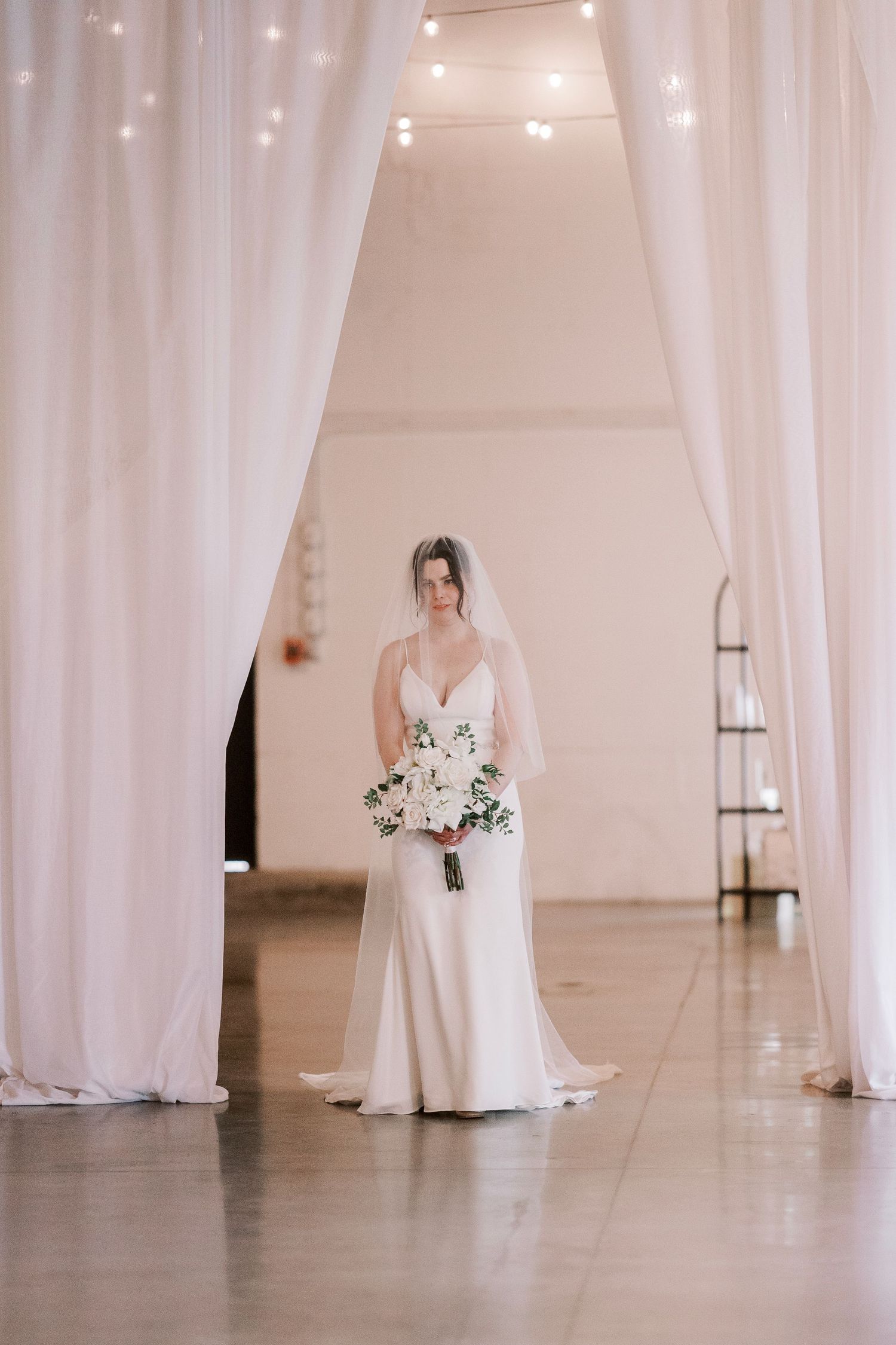 Bride in sleek white gown stands between draped white curtains holding bouquet.