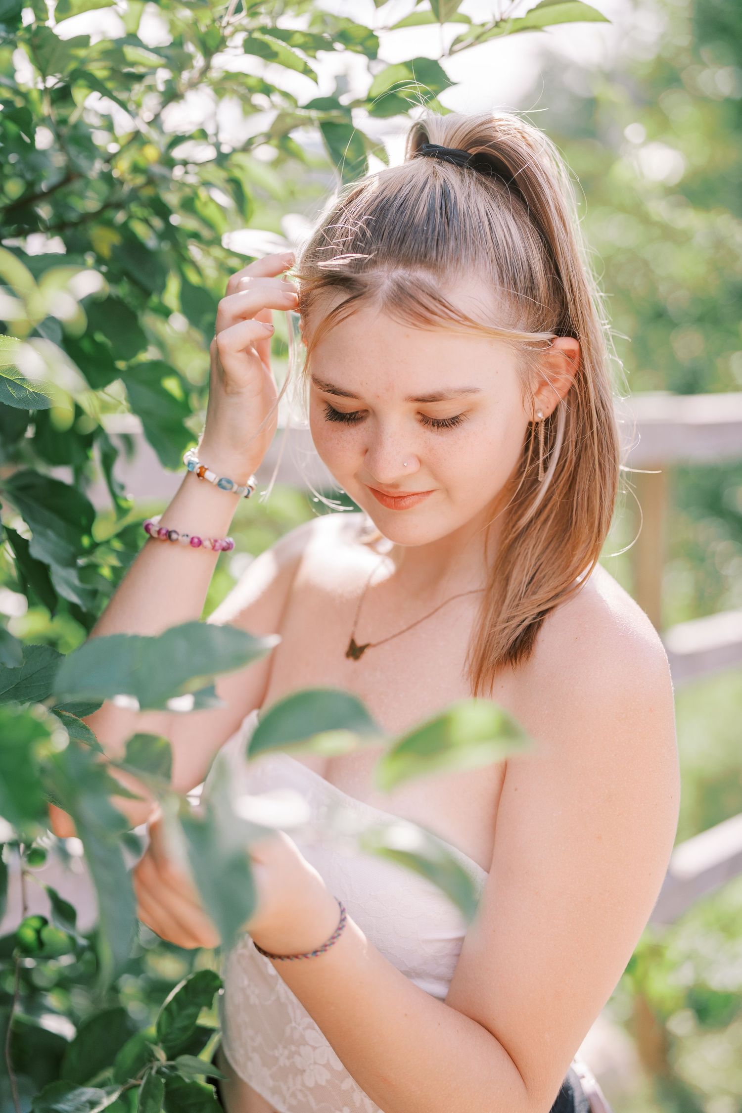 Ethereal portrait of woman in delicate dress standing among sun-dappled green foliage on a bright summer day.