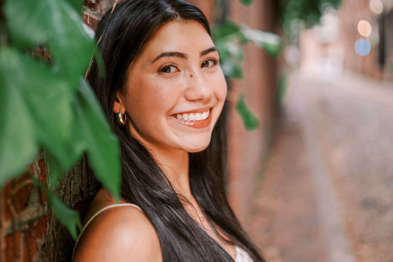 Portrait of a woman with long dark hair smiling against a brick wall with green ivy leaves in the background.