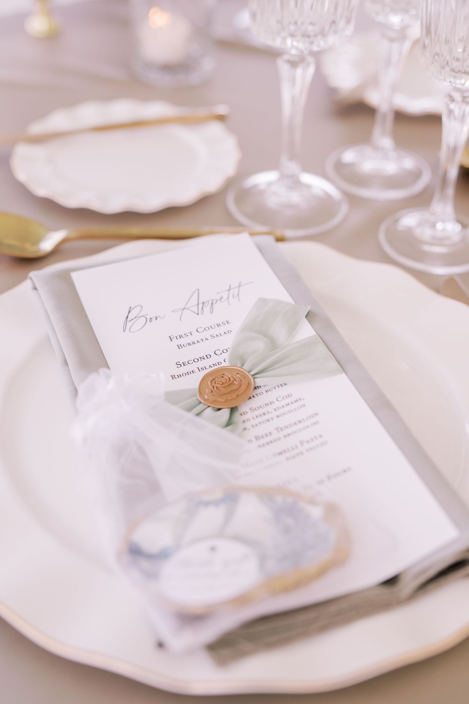 Elegant wedding menu card with gold wax seal placed on white plate with crystal wine glasses in background.