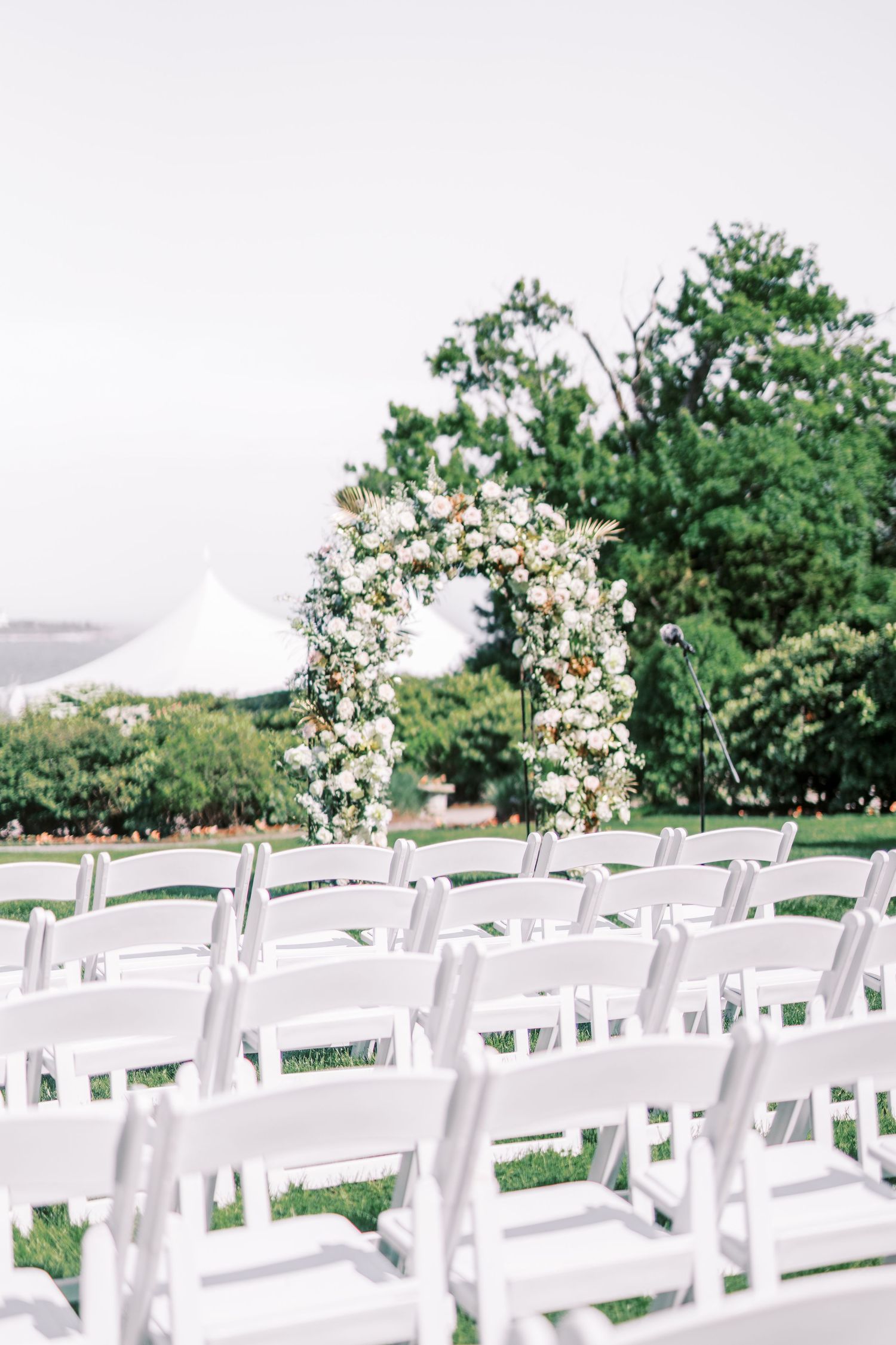 White folding chairs arranged in rows for an outdoor wedding ceremony with floral arrangements and trees in the background.