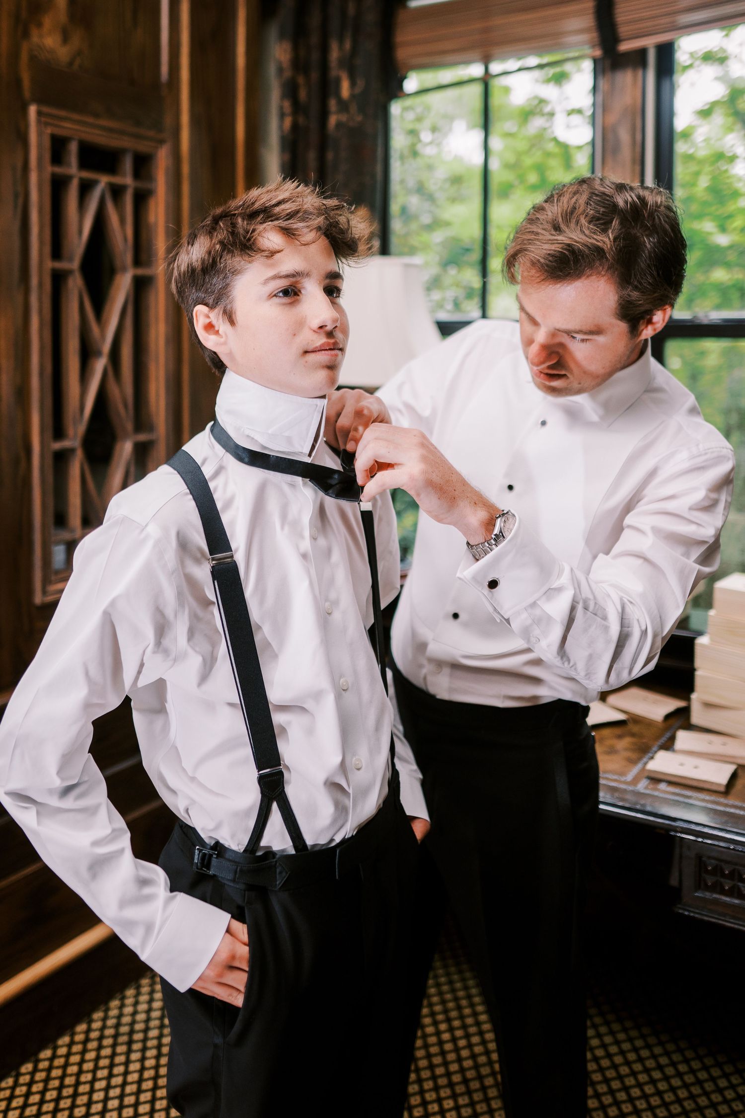 Two people in formal white shirts and suspenders help each other get dressed for a special occasion.