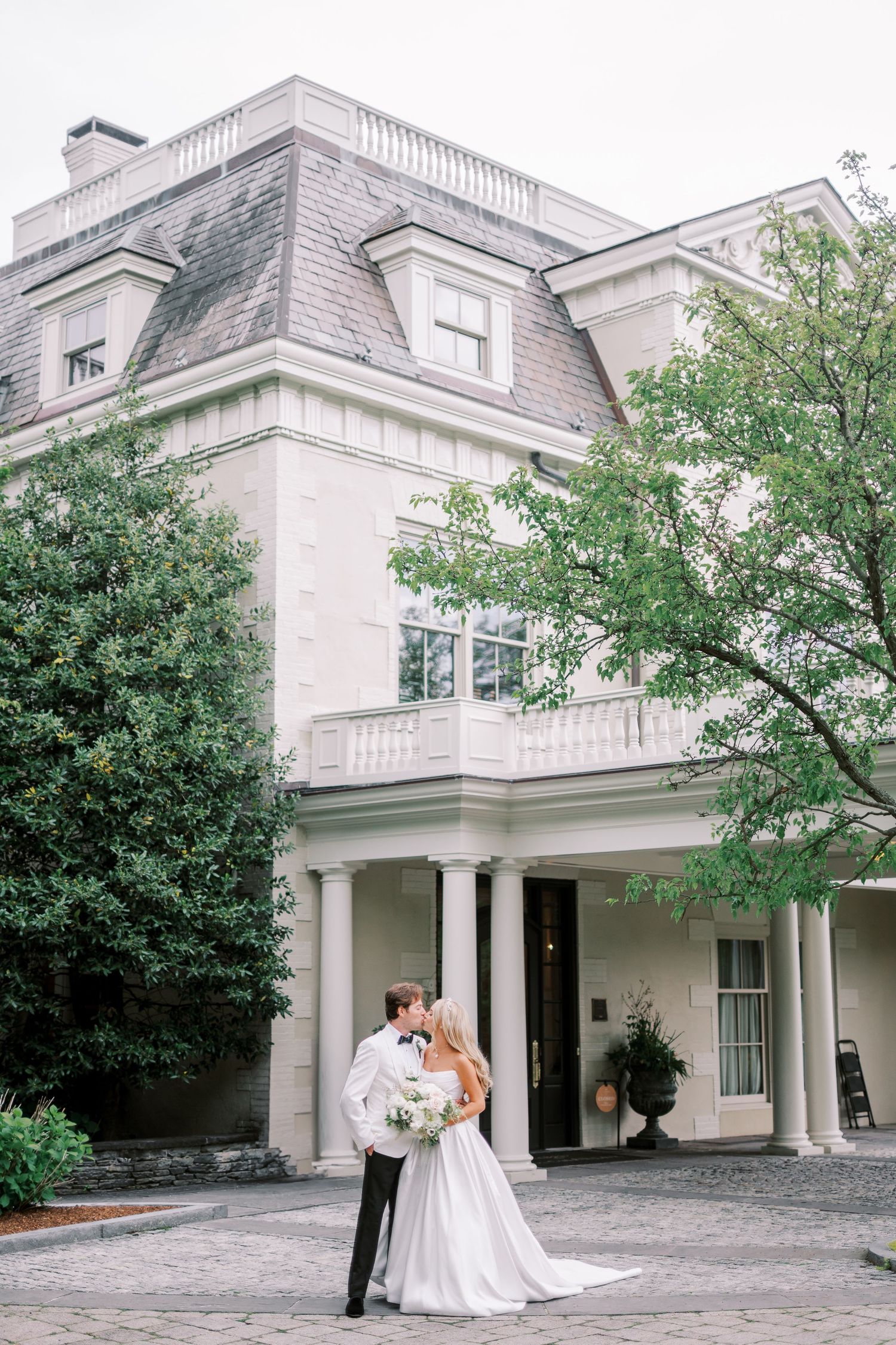 Classic white mansion with elegant columns and landscaping serves as backdrop for a romantic outdoor wedding ceremony.