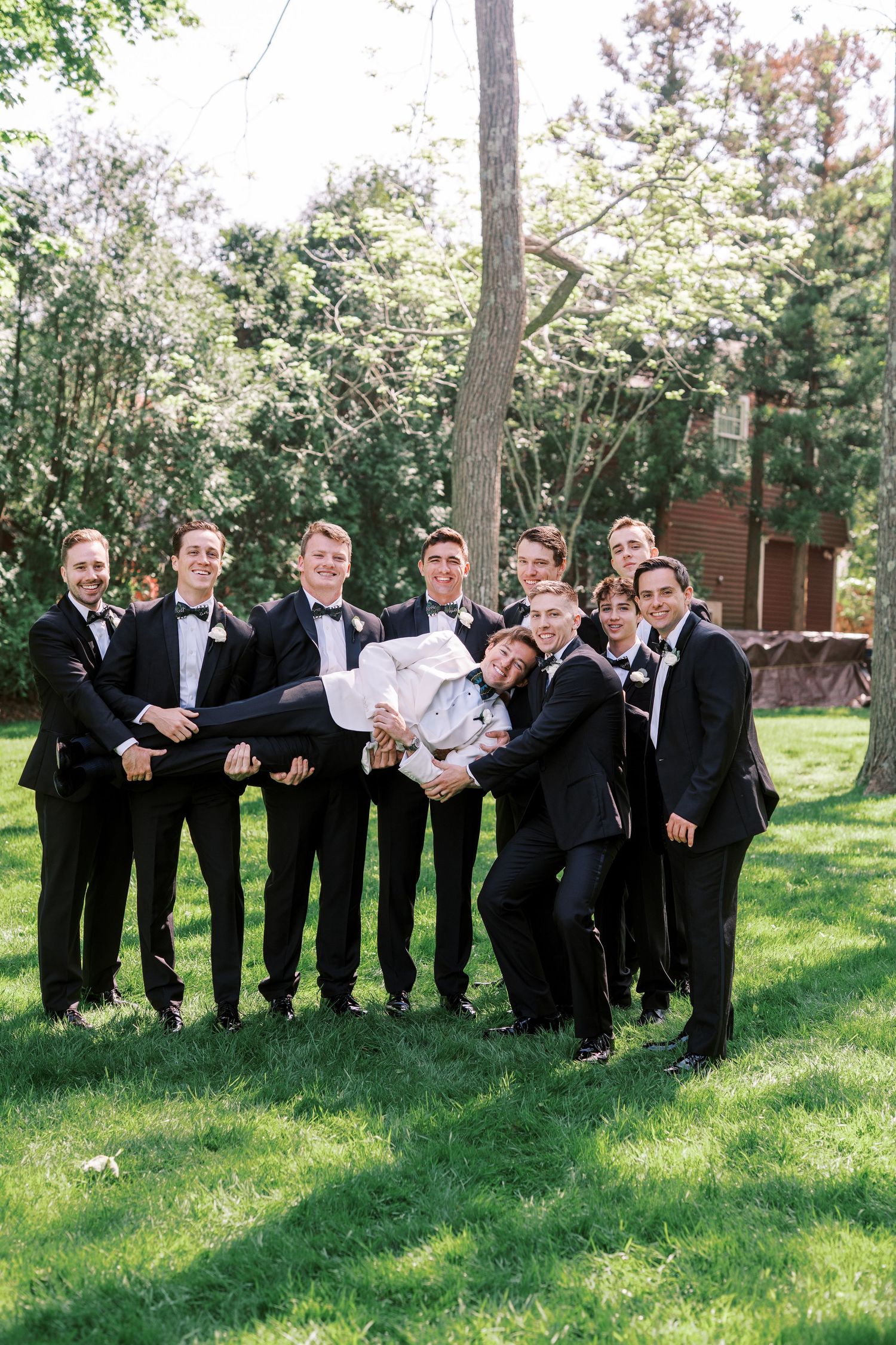 A group of groomsmen in black tuxedos pose playfully in a line on a sunny lawn with trees in the background.
