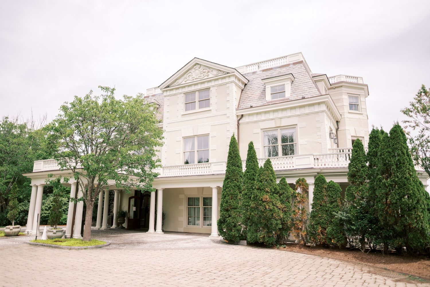 Elegant white mansion with columned portico and landscaped evergreen shrubs stands against an overcast sky.