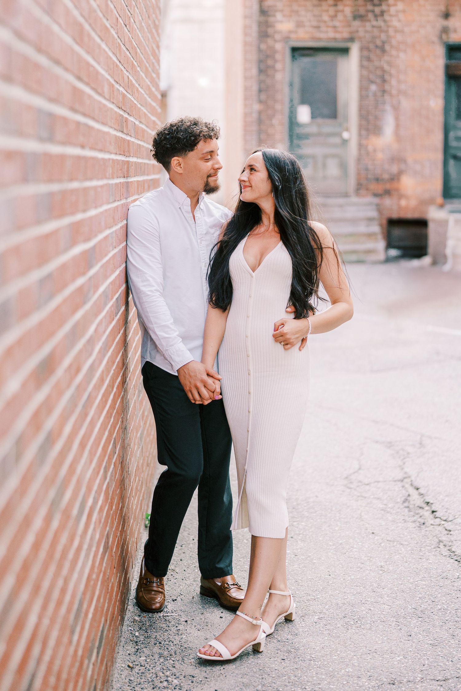 A romantic couple in white and black attire shares intimate moments against a rustic brick wall setting during engagement photos.