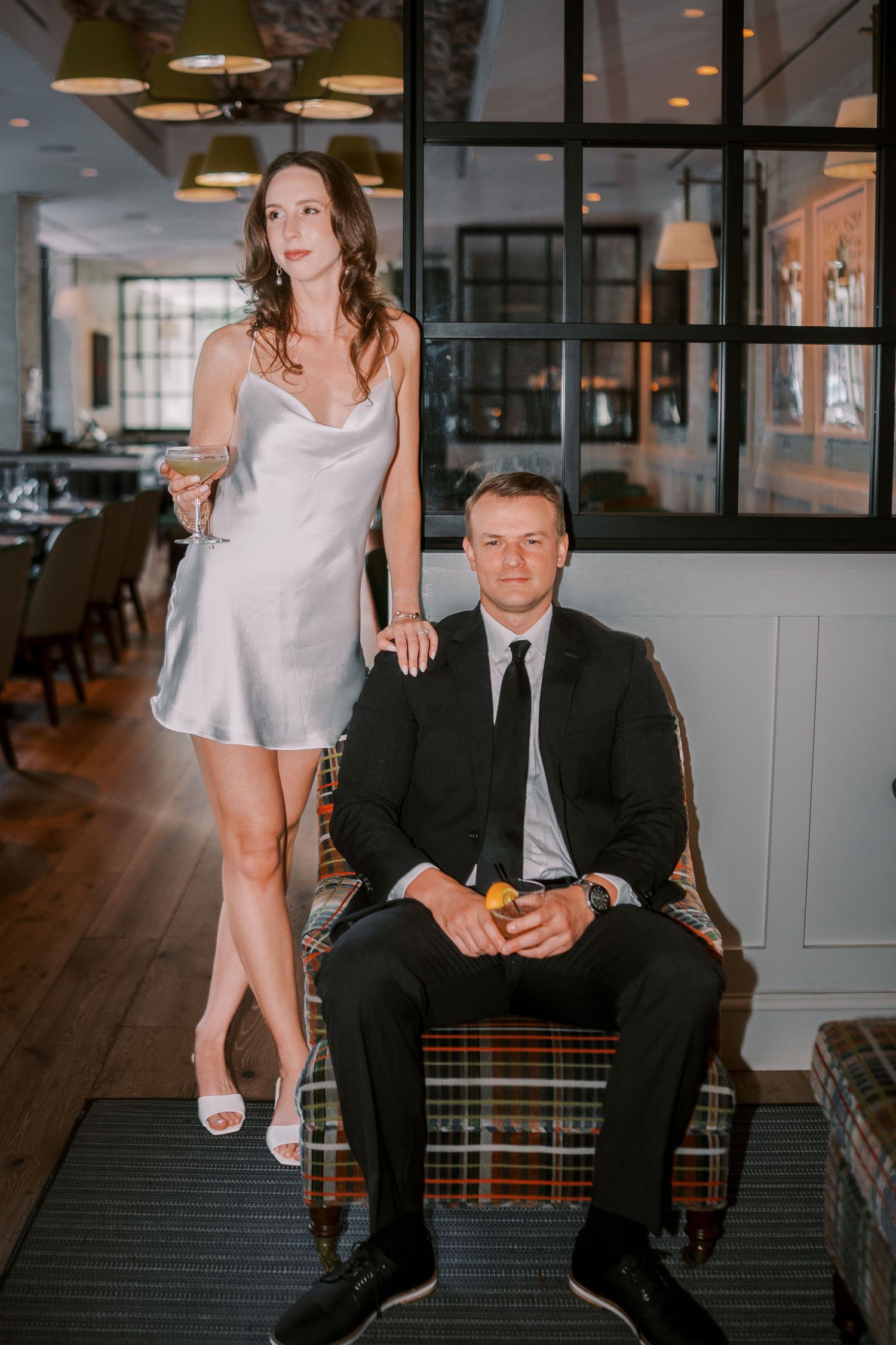 A couple poses in a modern restaurant interior with elegant decor, woman in silver dress and man in black suit.