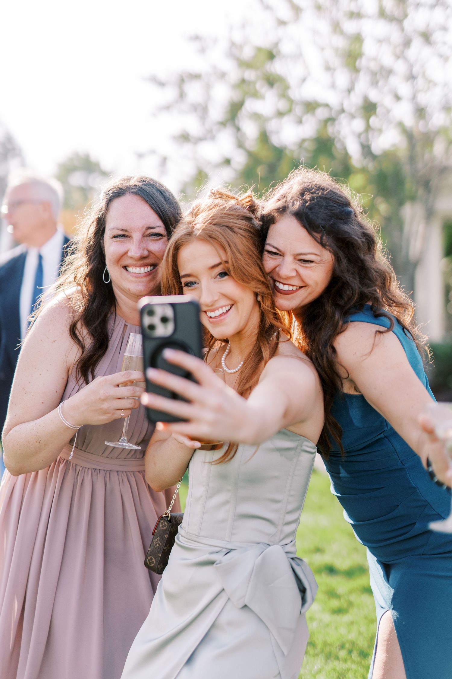 A joyful wedding party in coordinated blue and mauve dresses pose together outdoors for smartphone photos.