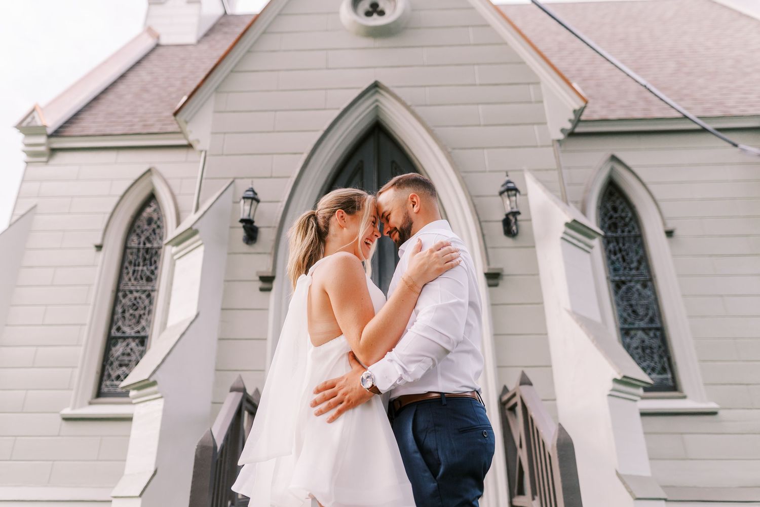 A couple shares an intimate moment outside a white church with gothic windows and lanterns.