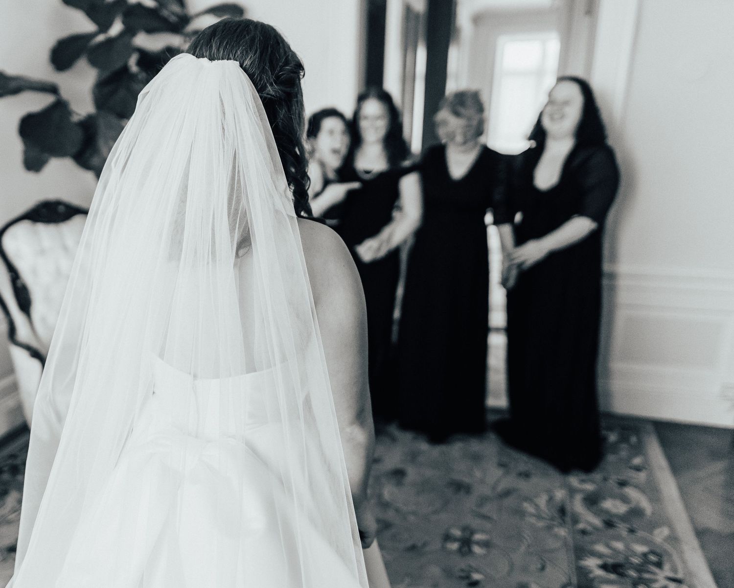 A bride in a white wedding dress and veil stands with bridesmaids wearing black dresses in an elegant room.