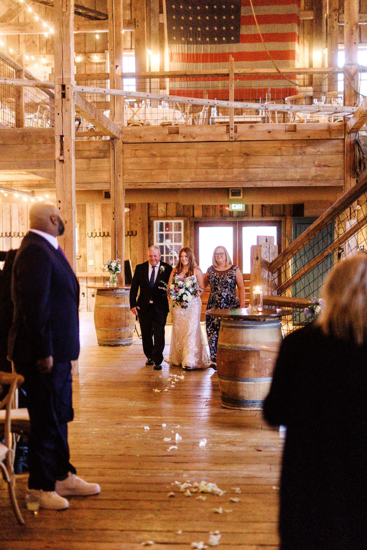 A rustic wedding processional walks down a wooden barn interior with American flag decor and wooden barrels.