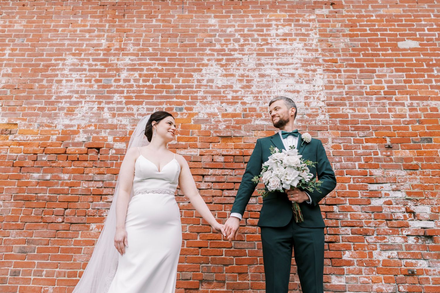 Bride and groom stand against weathered brick wall with white floral bouquet.