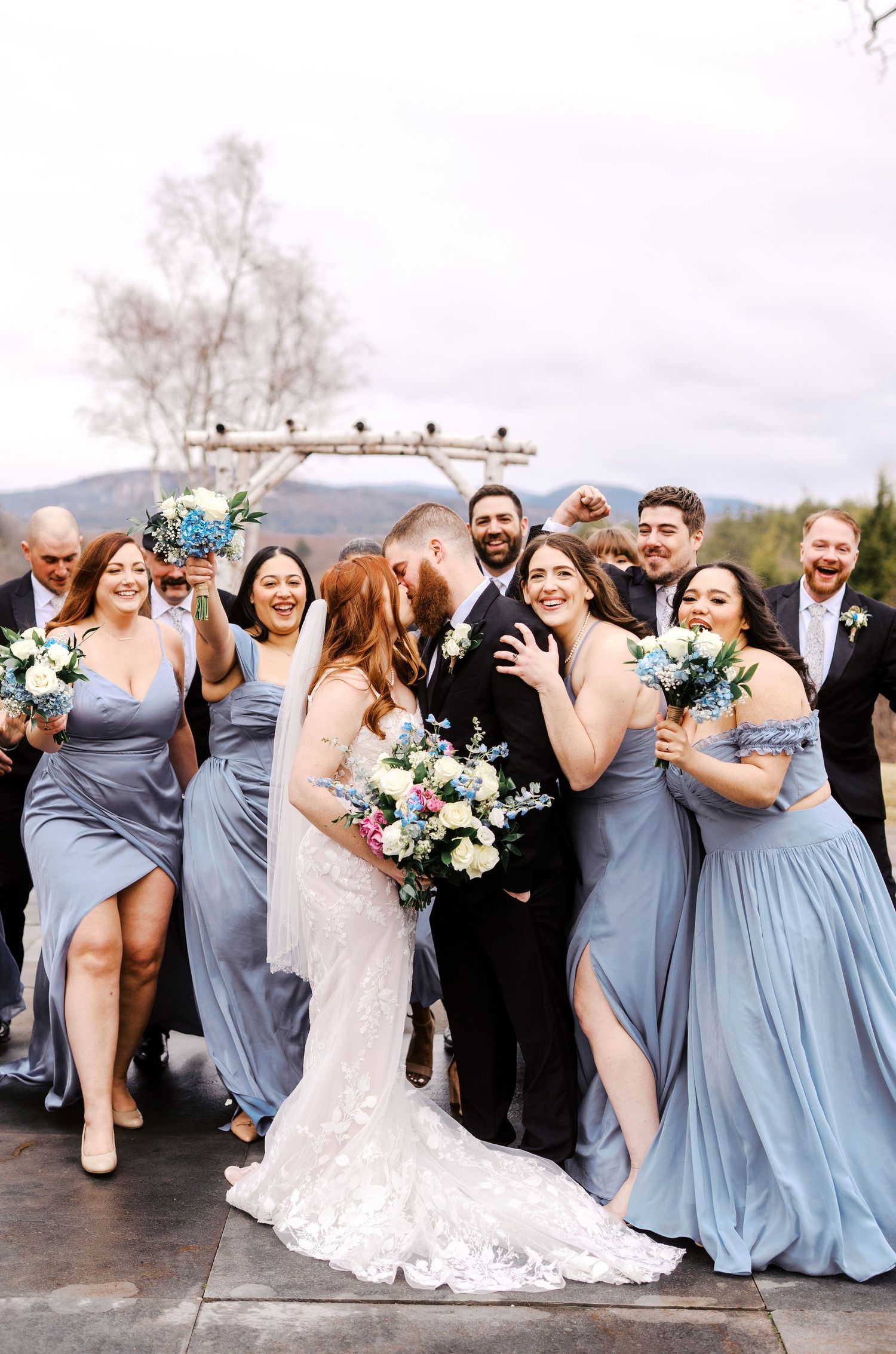 Wedding party in blue bridesmaid dresses laugh joyfully with bride in white lace gown during outdoor celebration.