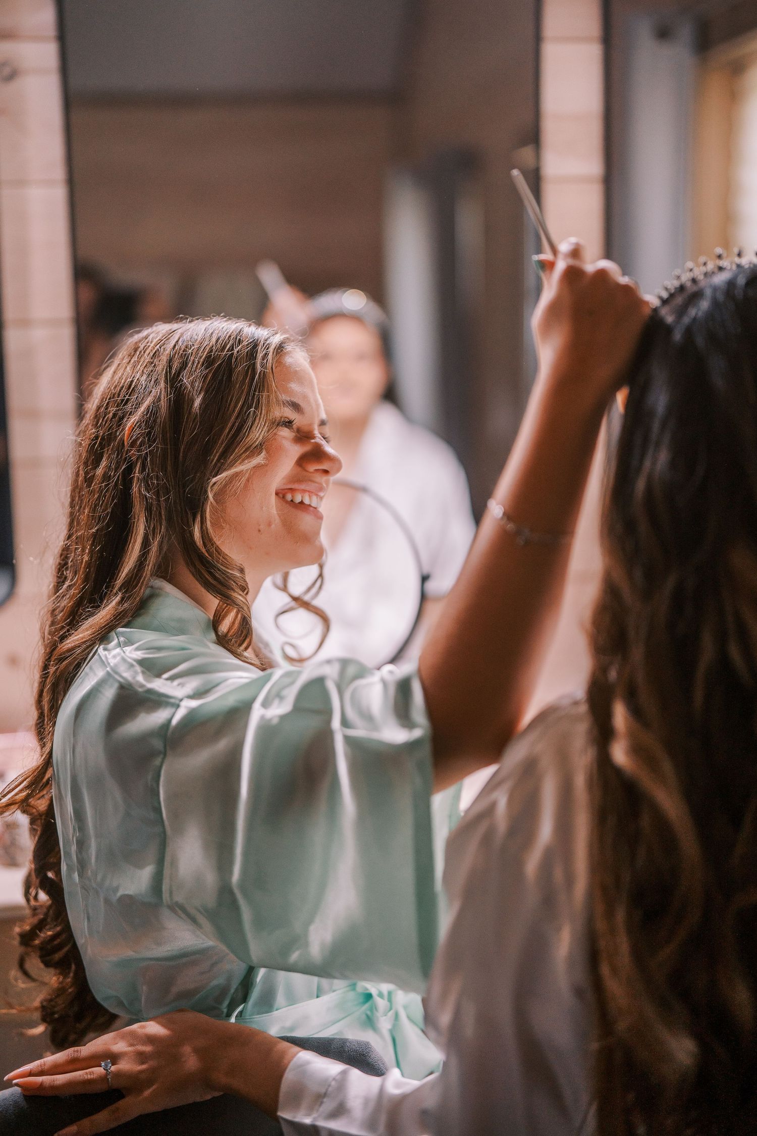 A woman in a light satin robe laughs joyfully while getting ready, with soft natural lighting in the background.