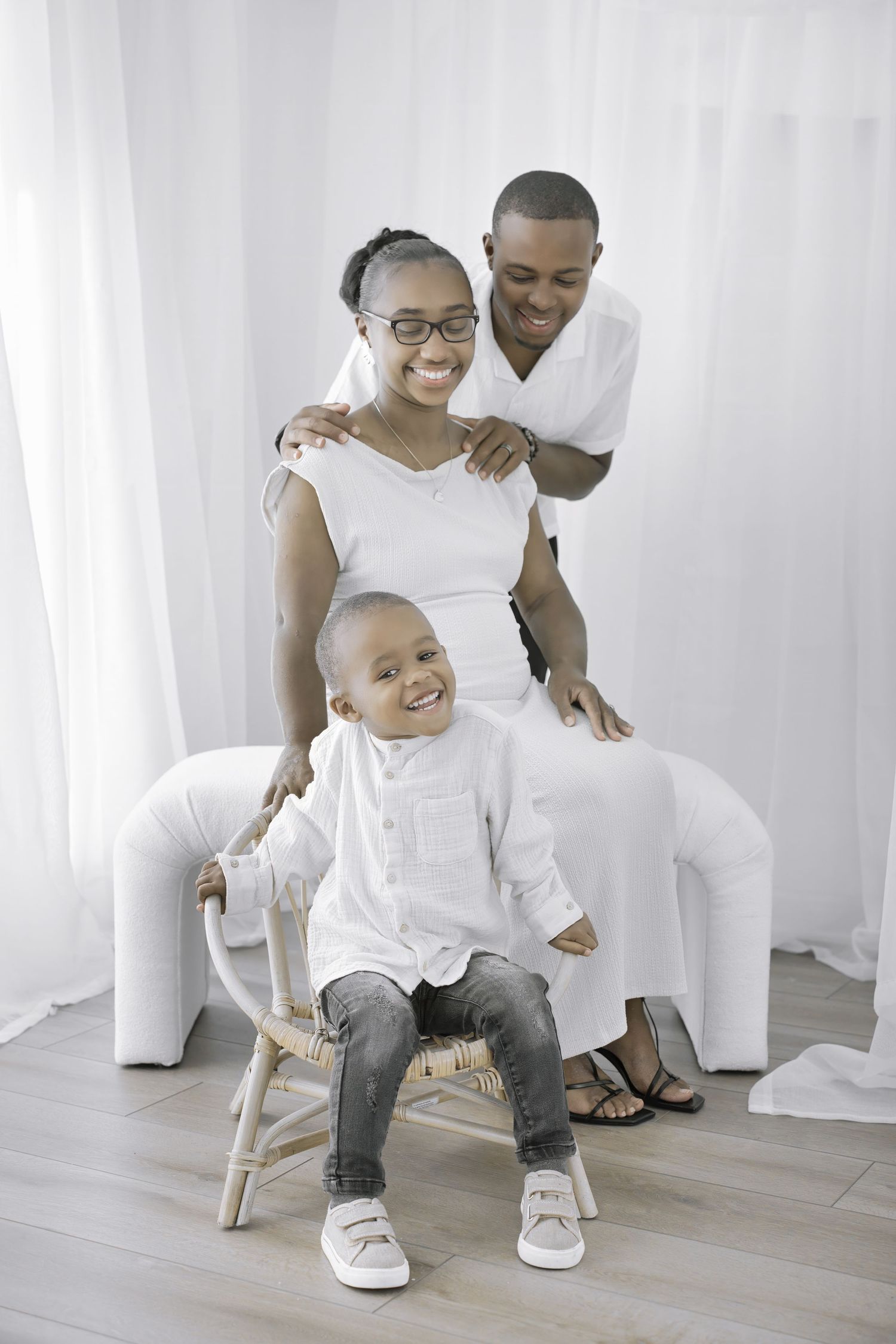 Portrait of a joyful family in white outfits sitting together on a chair against a light background.