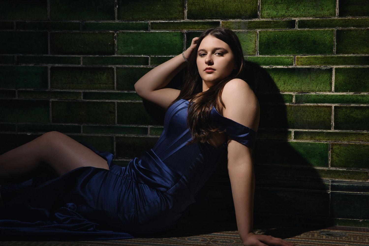 Dramatic portrait of a model in a navy blue evening gown posed against moss-covered brick walls.