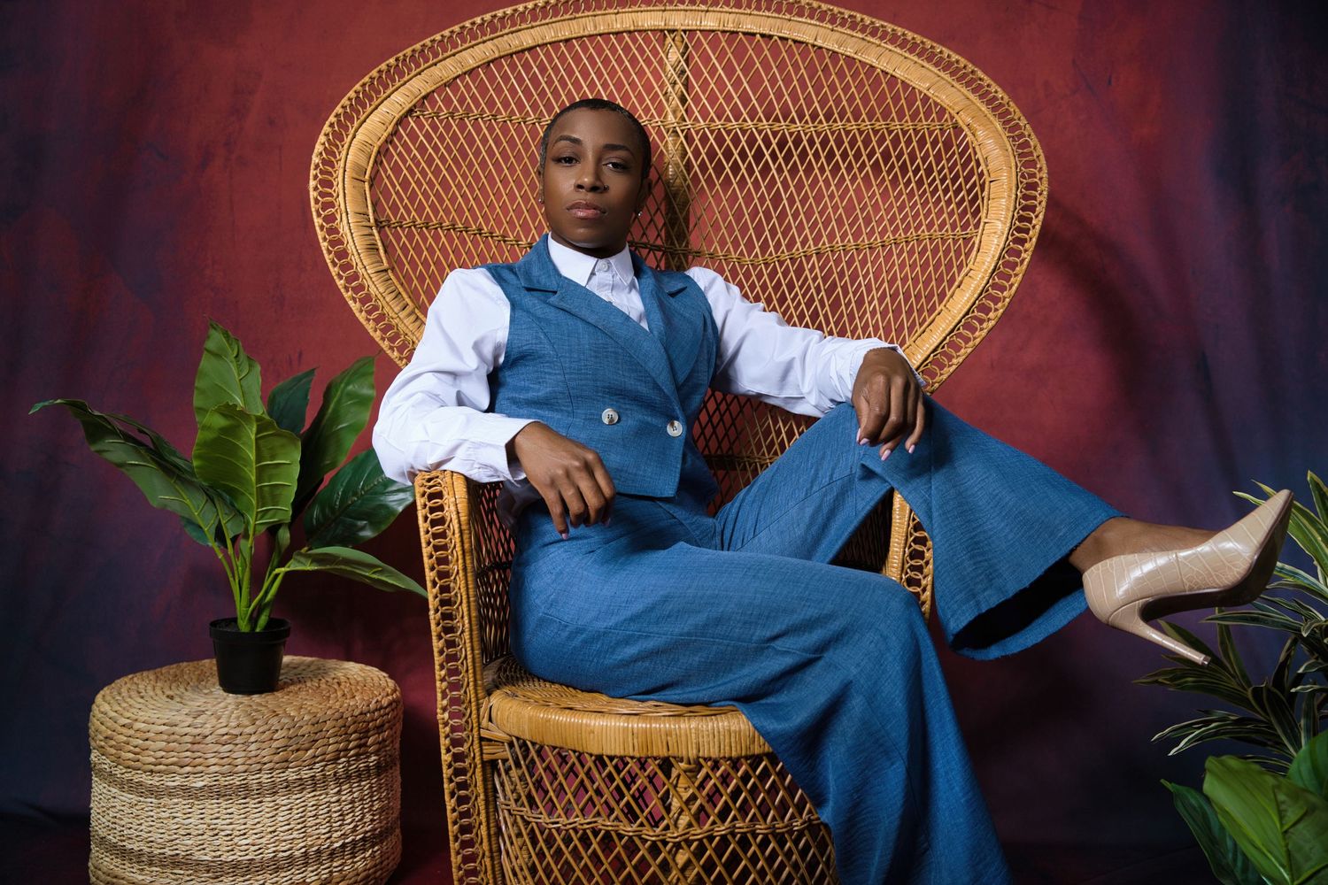Person in blue denim outfit sitting regally on a wicker peacock chair with plants in background.