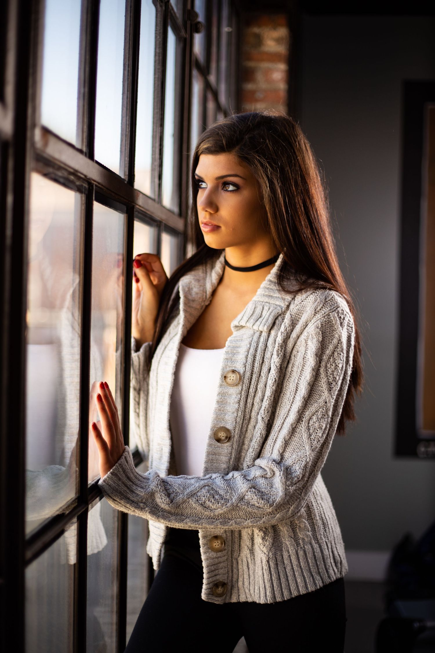 Thoughtful portrait by window of person wearing knit cardigan and white top with choker necklace.