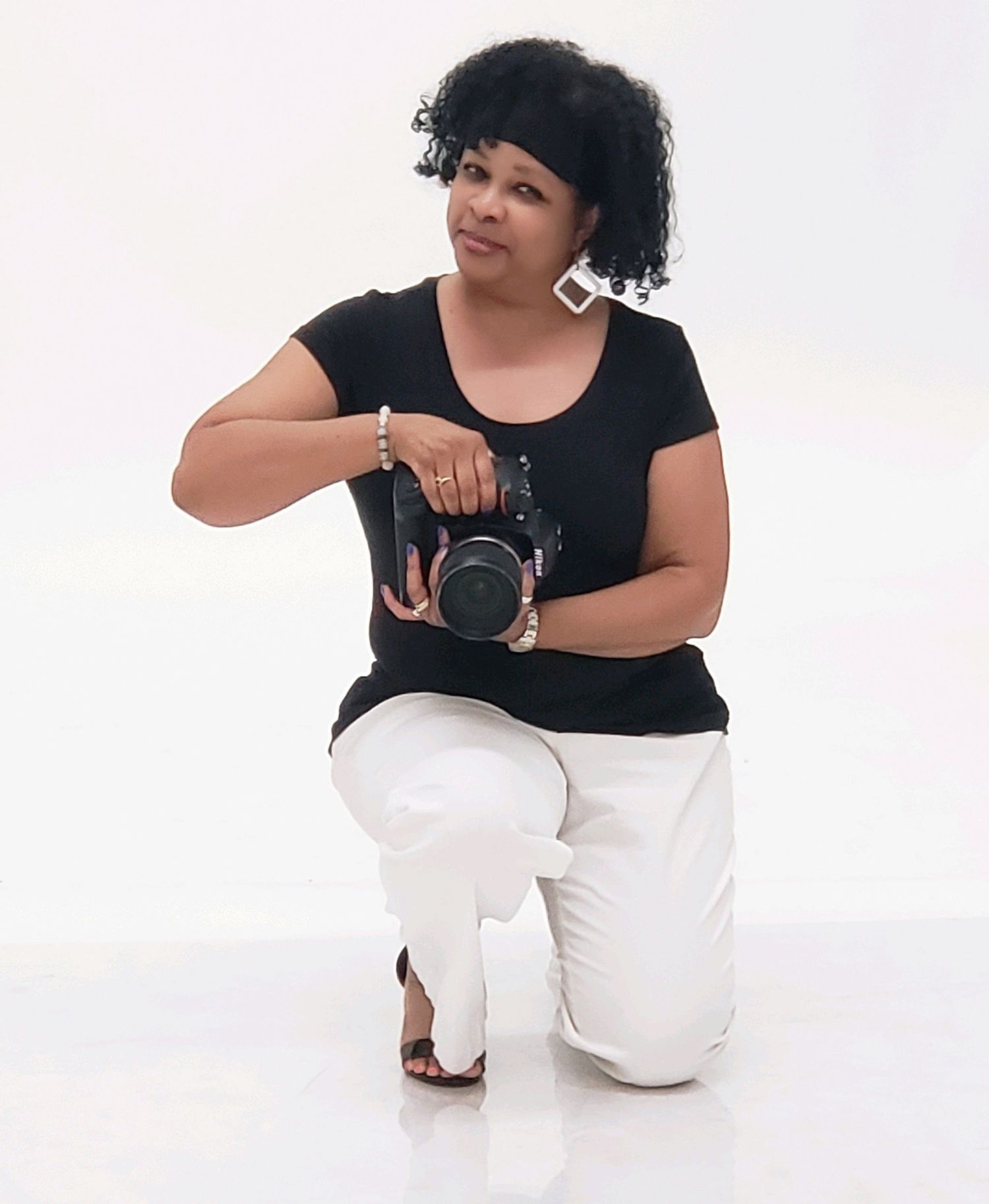 A photographer poses with a camera while kneeling in a studio against a white background.