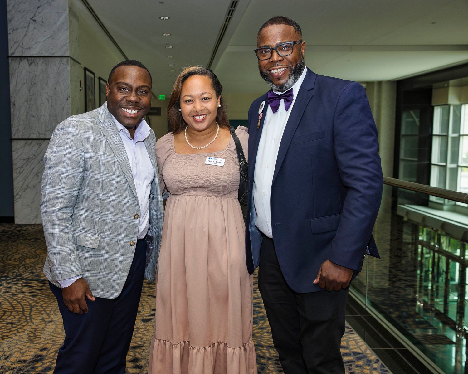 Three elegantly dressed people pose together at a formal event in front of a marble wall.