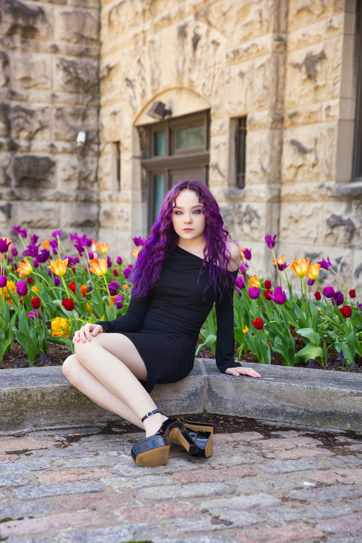 Woman in black dress with vibrant purple hair posing by tulip garden and stone architecture.