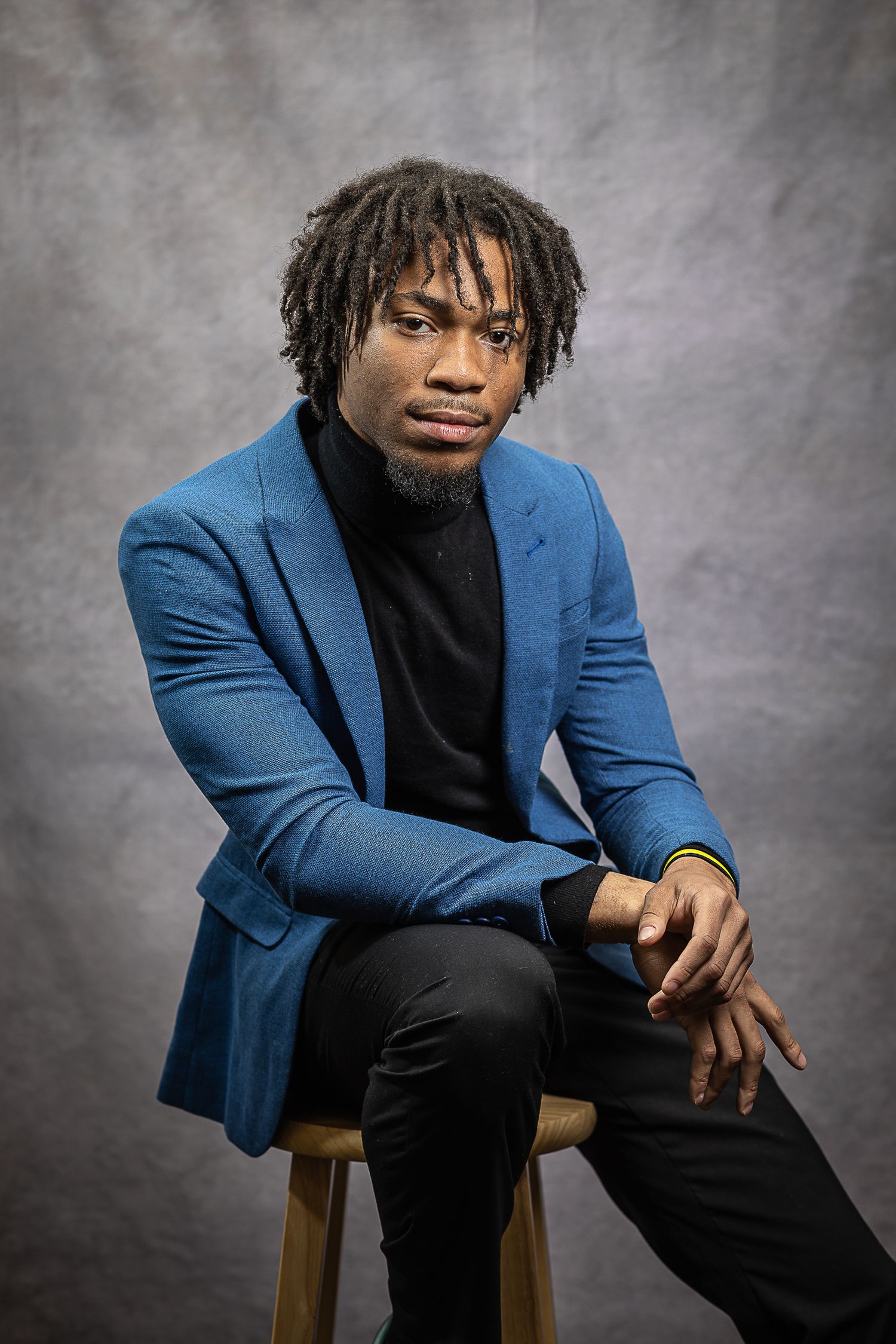 Professional studio portrait featuring a person in a blue blazer and black turtleneck seated on a wooden stool.