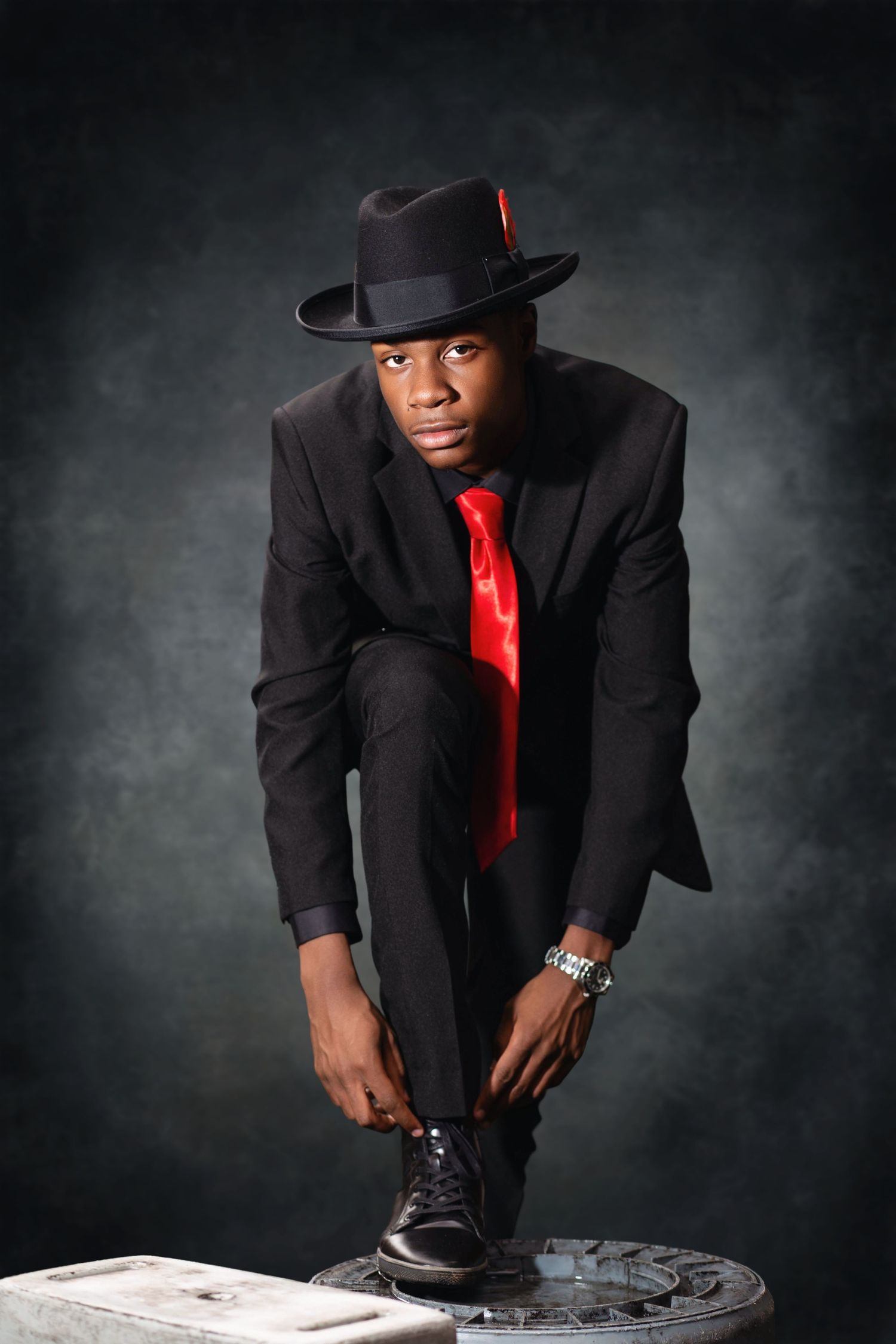 Stylish portrait of person in black suit, red tie and fedora posing dramatically on white platform.
