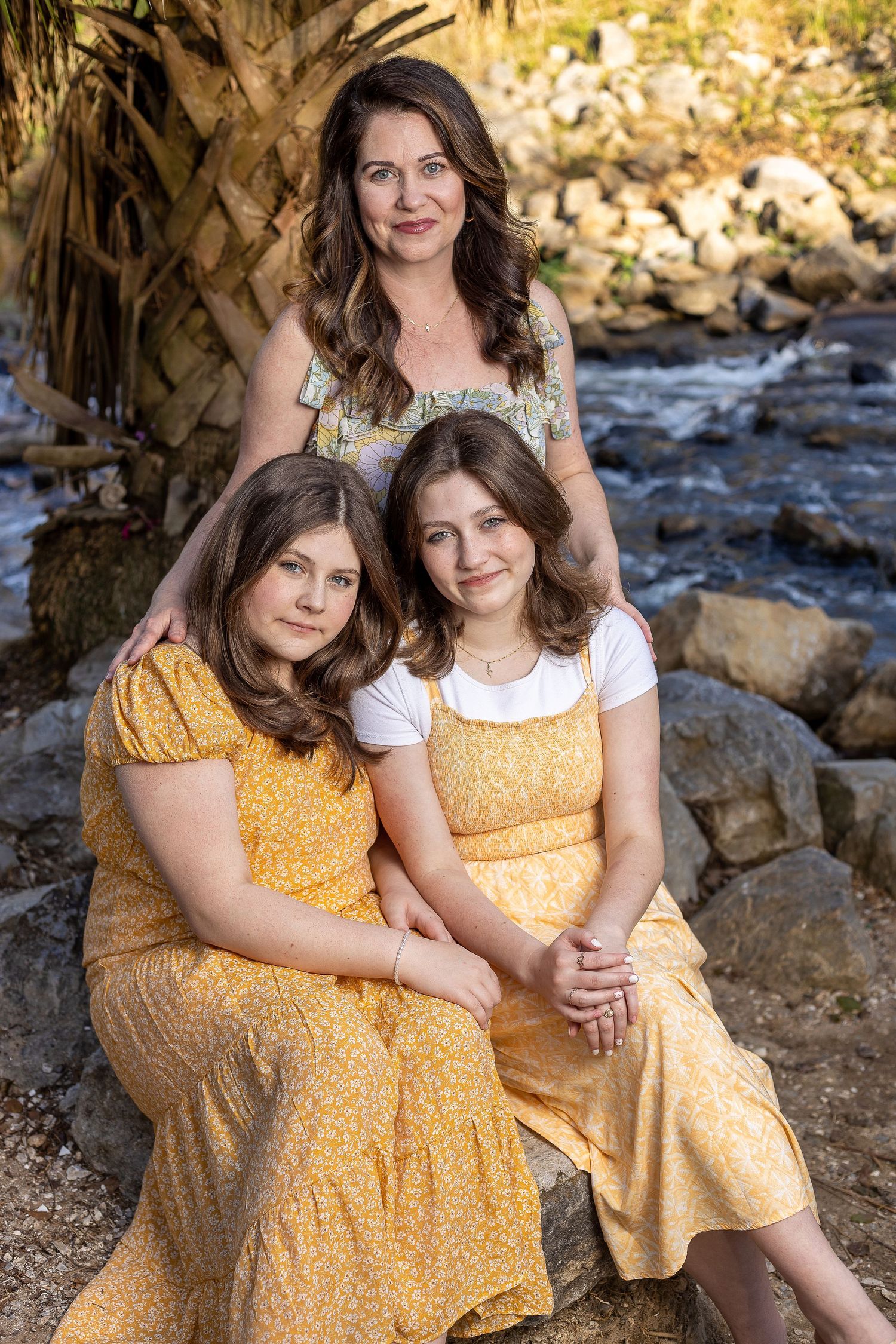 Family portrait of three people in yellow and white dresses posed by a scenic stream.
