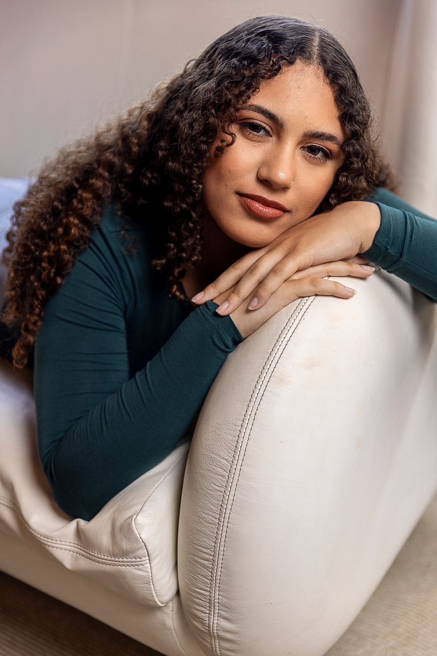 Professional portrait of a person with curly hair in a green top leaning on white furniture.