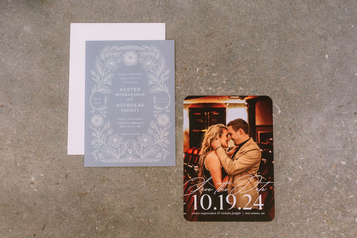 Elegant wedding invitation suite in dusty blue and gray tones with floral designs and multiple coordinating cards spread on concrete.