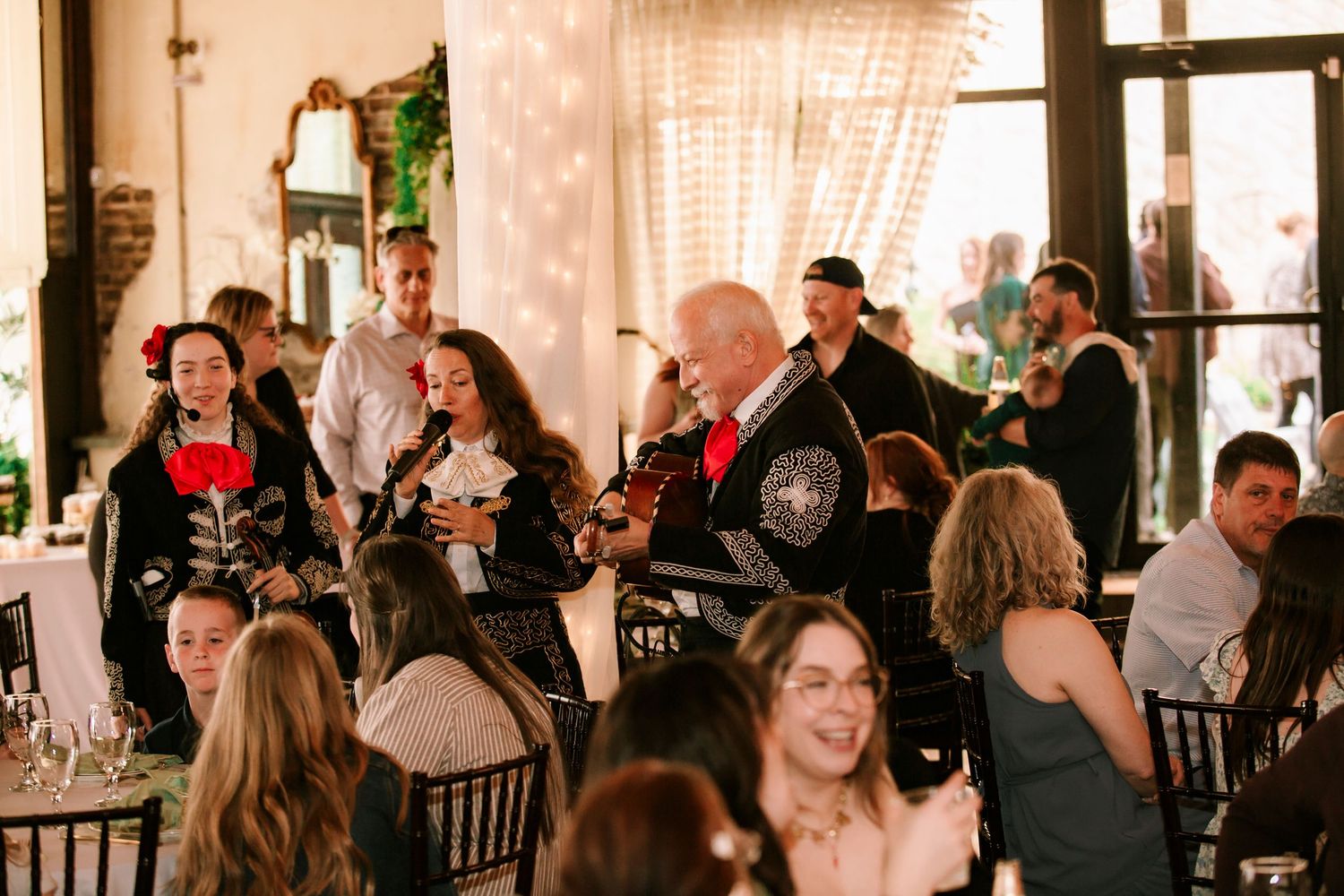 A lively wedding reception party with guests mingling at tables and entertainers performing folk music in a rustic venue.