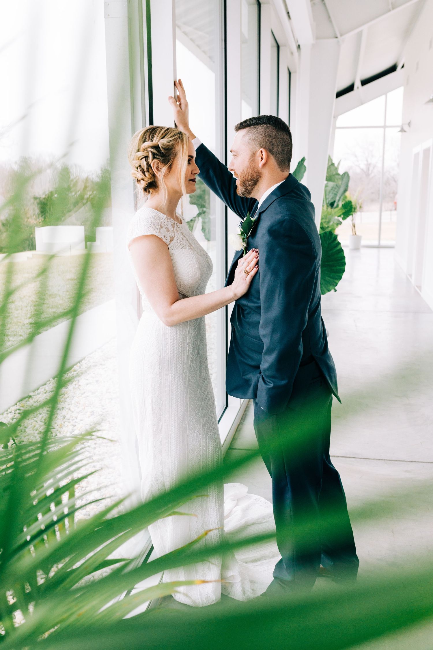 A bride and groom share an intimate moment in a bright white hallway with tropical plants framing the scene at Greenhouse Two Rivers in MO