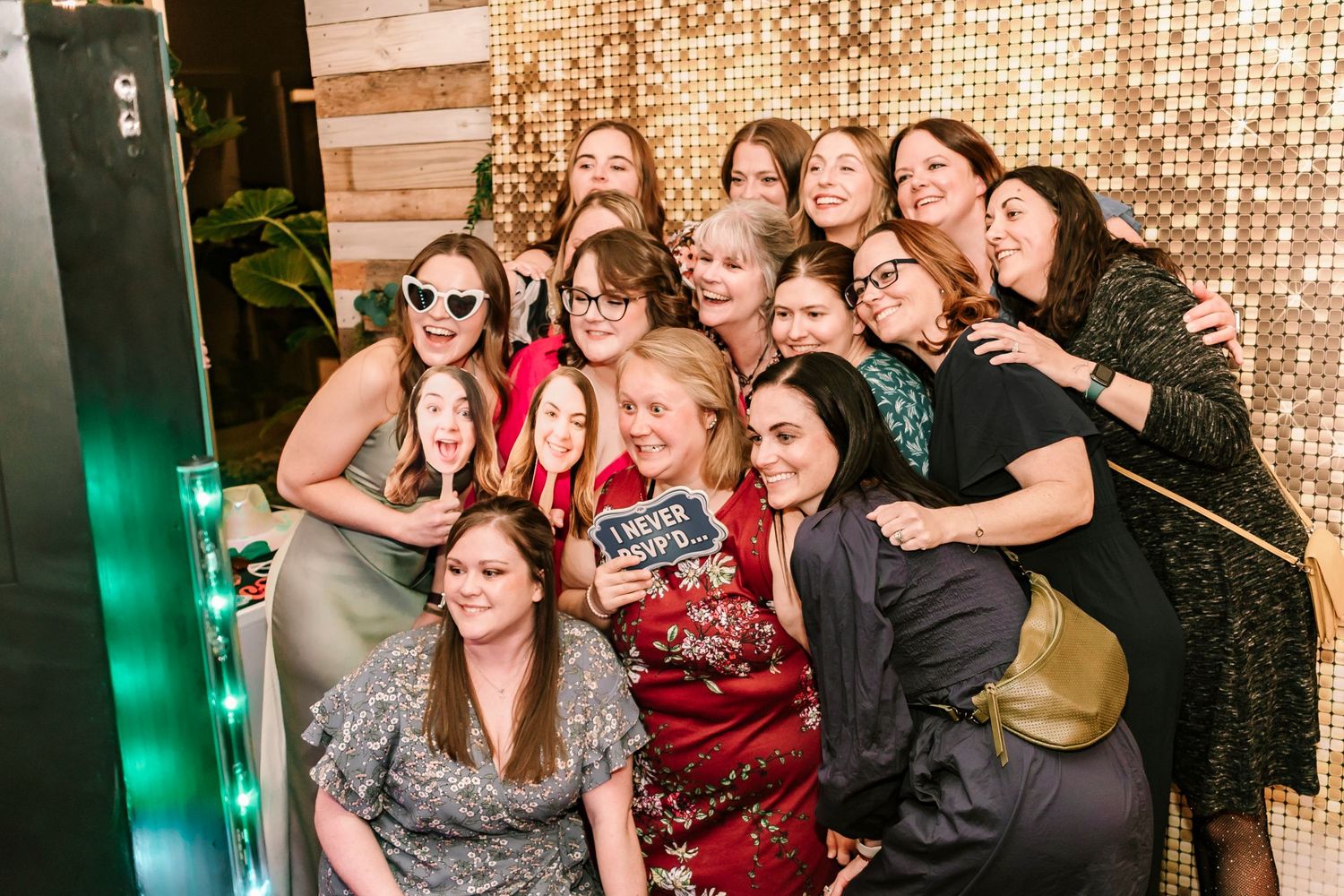 A fun group photo at a party with people posing together in front of a sparkly gold sequined wall with green lighting.