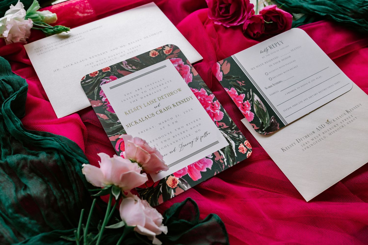 Wedding stationery with floral designs laid out on deep red velvet fabric with pink and red roses as decoration.