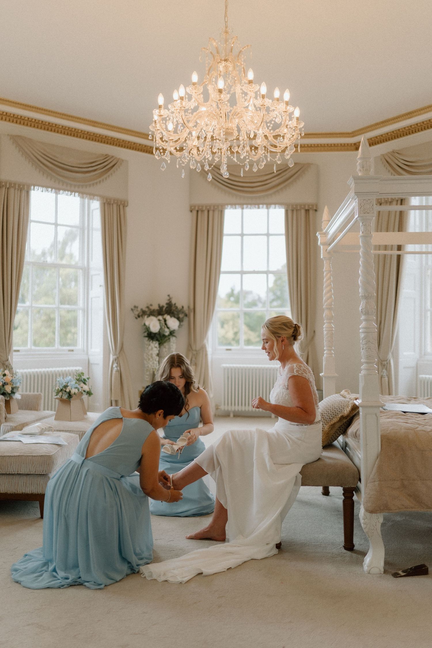 Elegant bridal preparation scene in a grand room with crystal chandelier and draped windows at Bourton Hall.