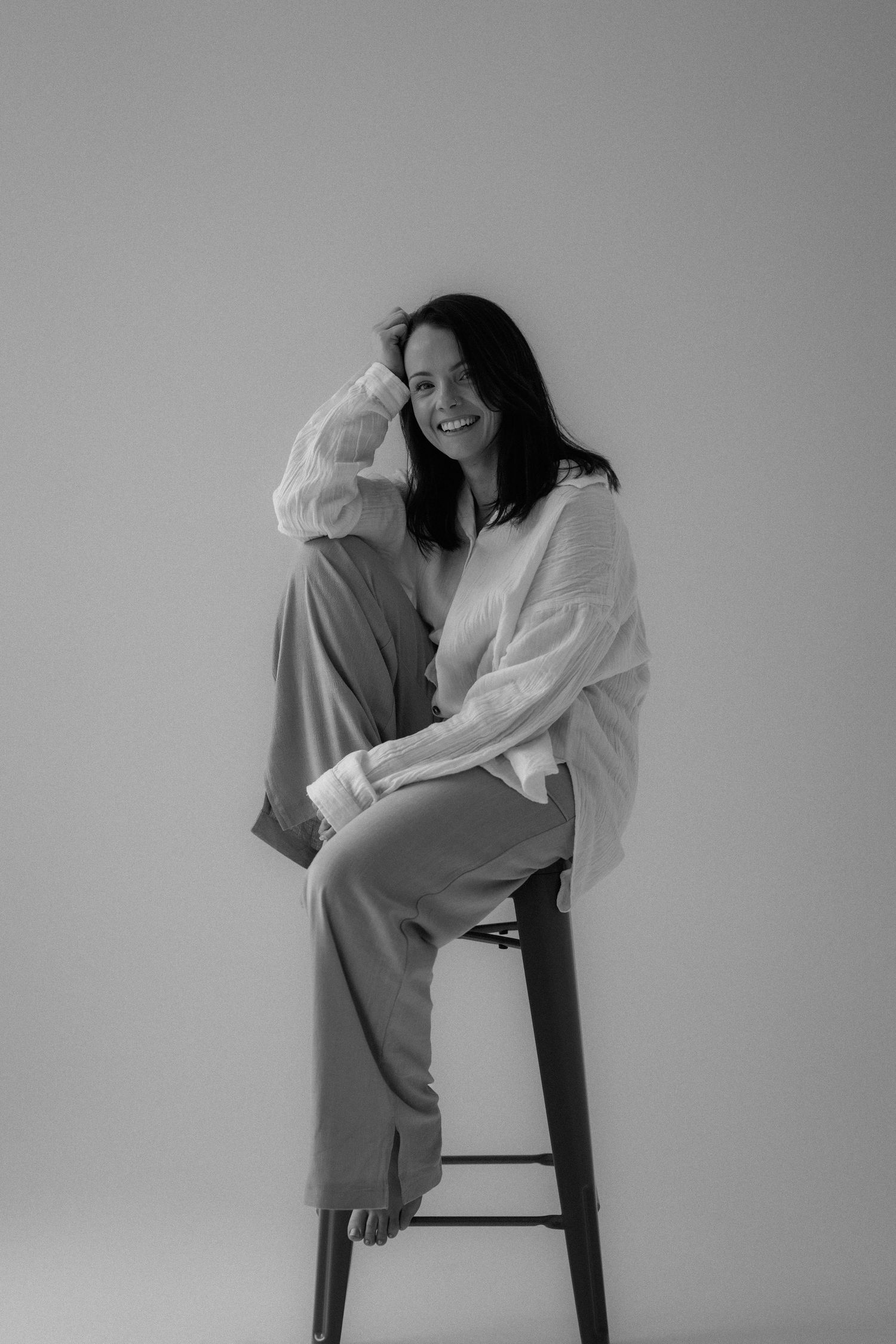 Black and white portrait of someone sitting casually on a bar stool wearing a white sweater and grey pants.