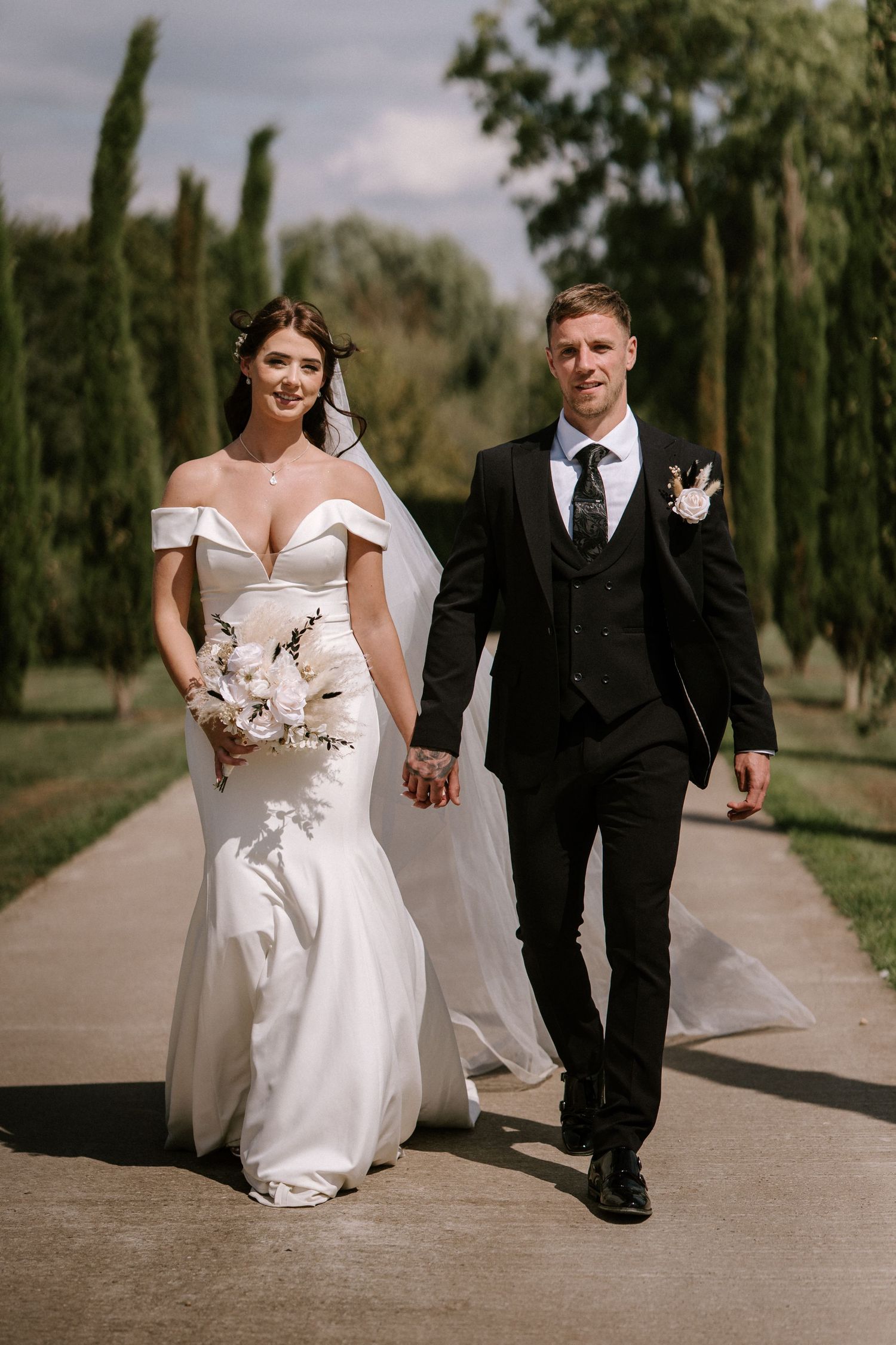 Newlywed couple strolls along a cypress tree-lined path in an outdoor wedding setting. Wedding photography at Willow Marsh Farm.
