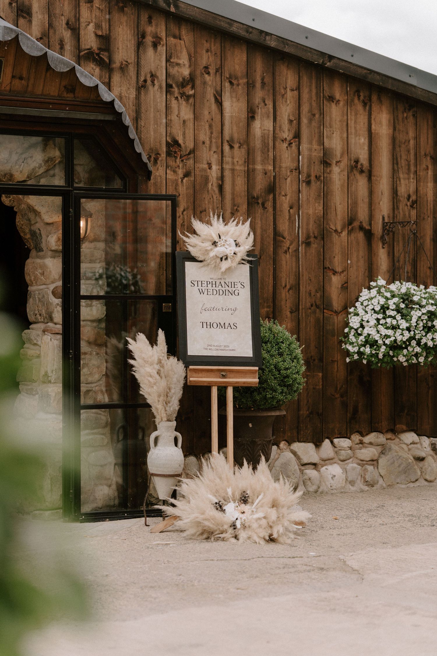 A rustic wooden barn entrance decorated with pampas grass and a wedding welcome sign on an stand. Wedding photography at Willow Marsh Farm.