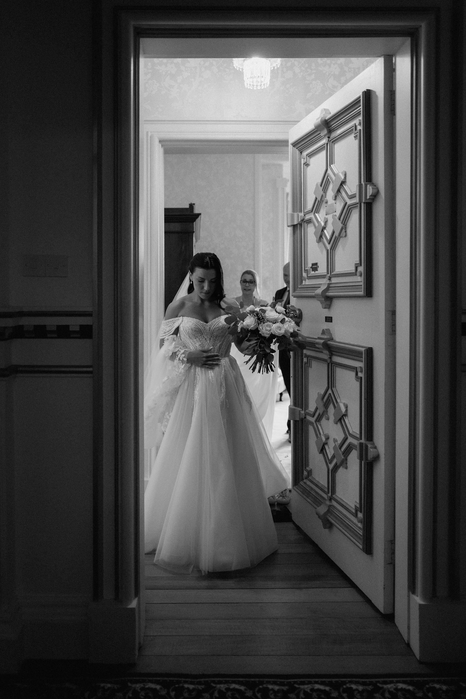 Black and white photo of bride with bouquet entering through ornate doorway at Harlaxton Manor.