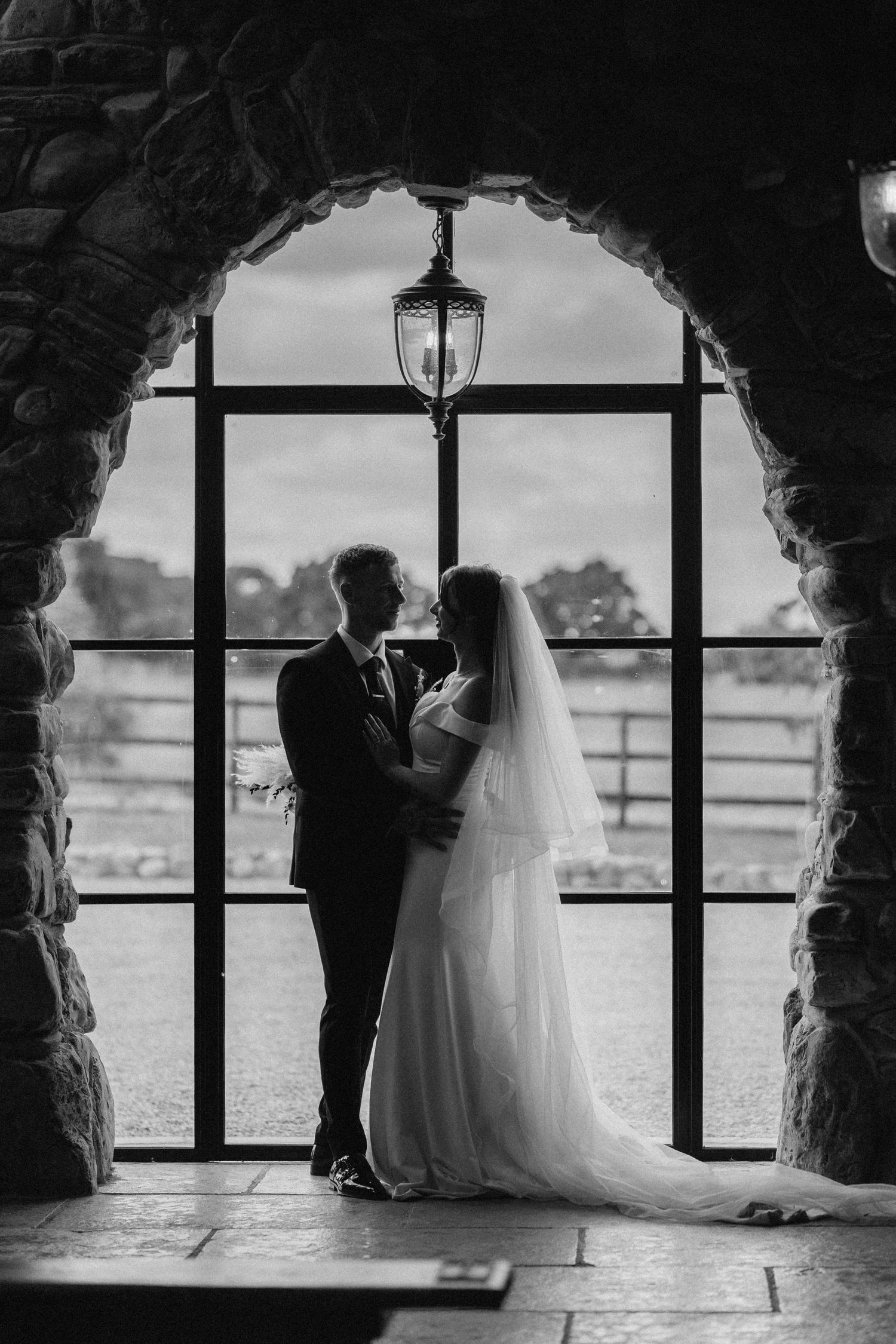 A romantic silhouette of a couple in an arched stone window frames a scenic waterfront view. Wedding photography at Willow Marsh Farm.