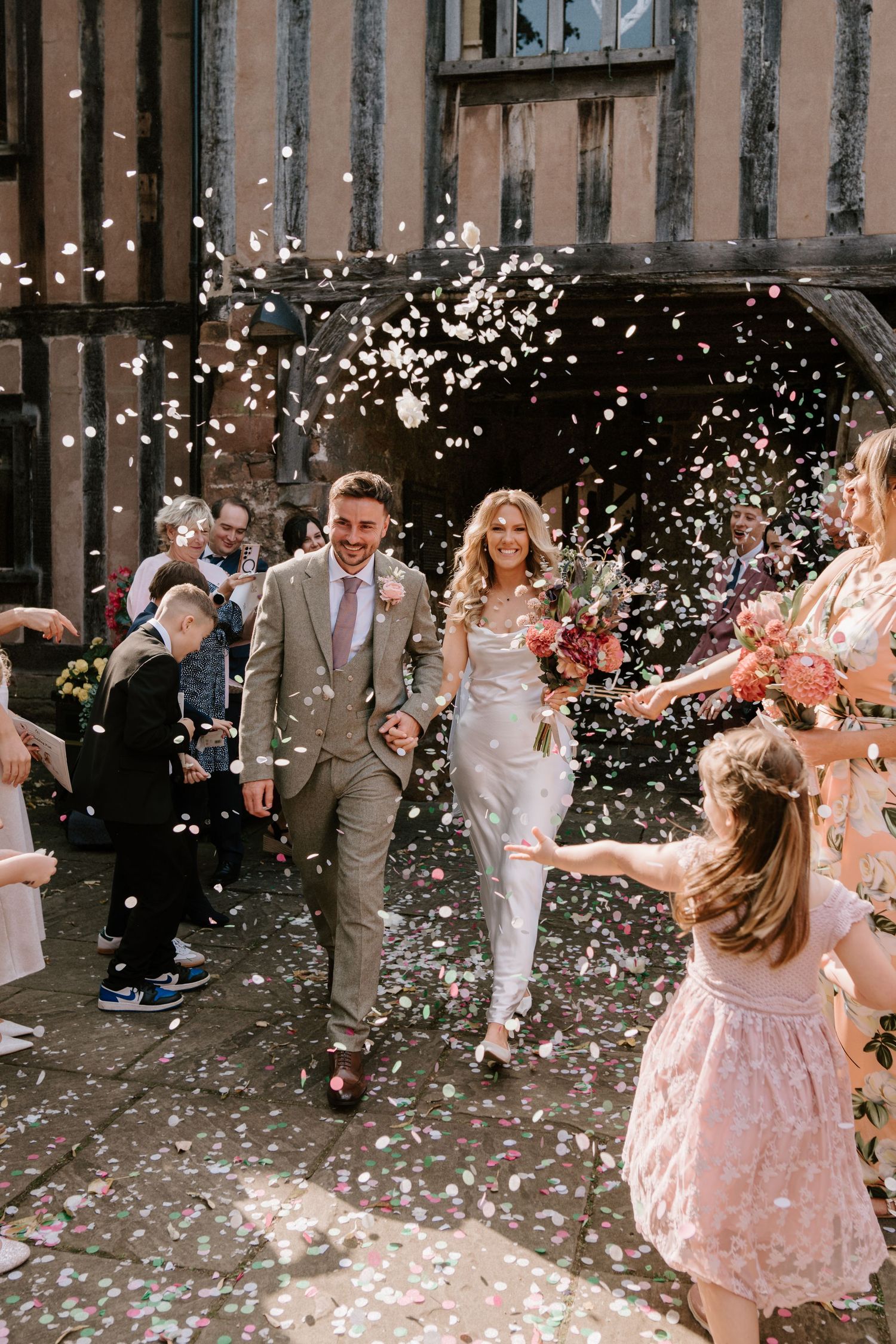 A couple walks through a shower of white petals and confetti as guests celebrate outside a rustic wooden building.