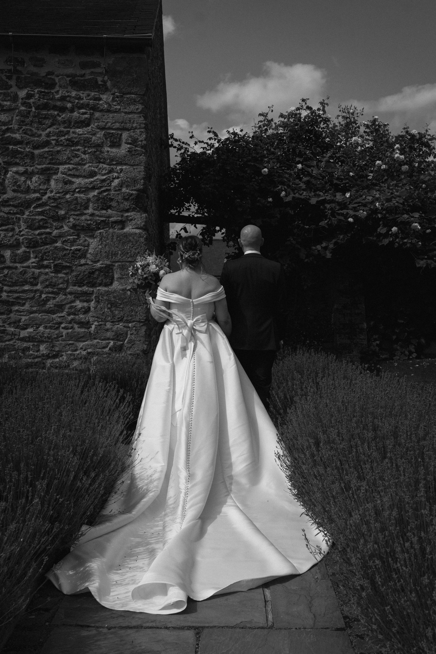 Black and white photo of a couple in formal attire walking along a stone wall with lavender at Dodford Manor.
